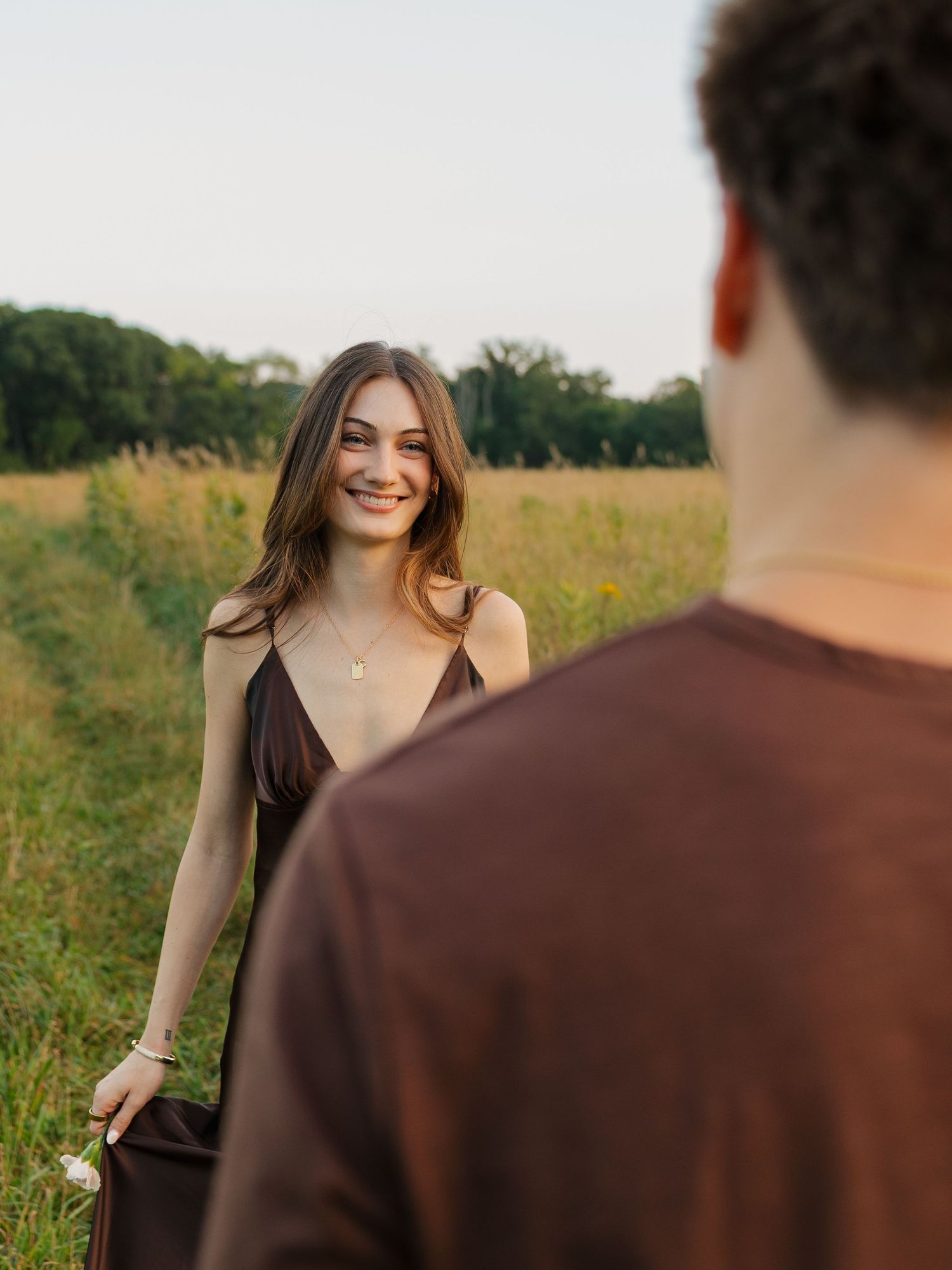 A smiling young woman in a brown dress stands in a sunlit field, looking back at a man in a brown shirt.