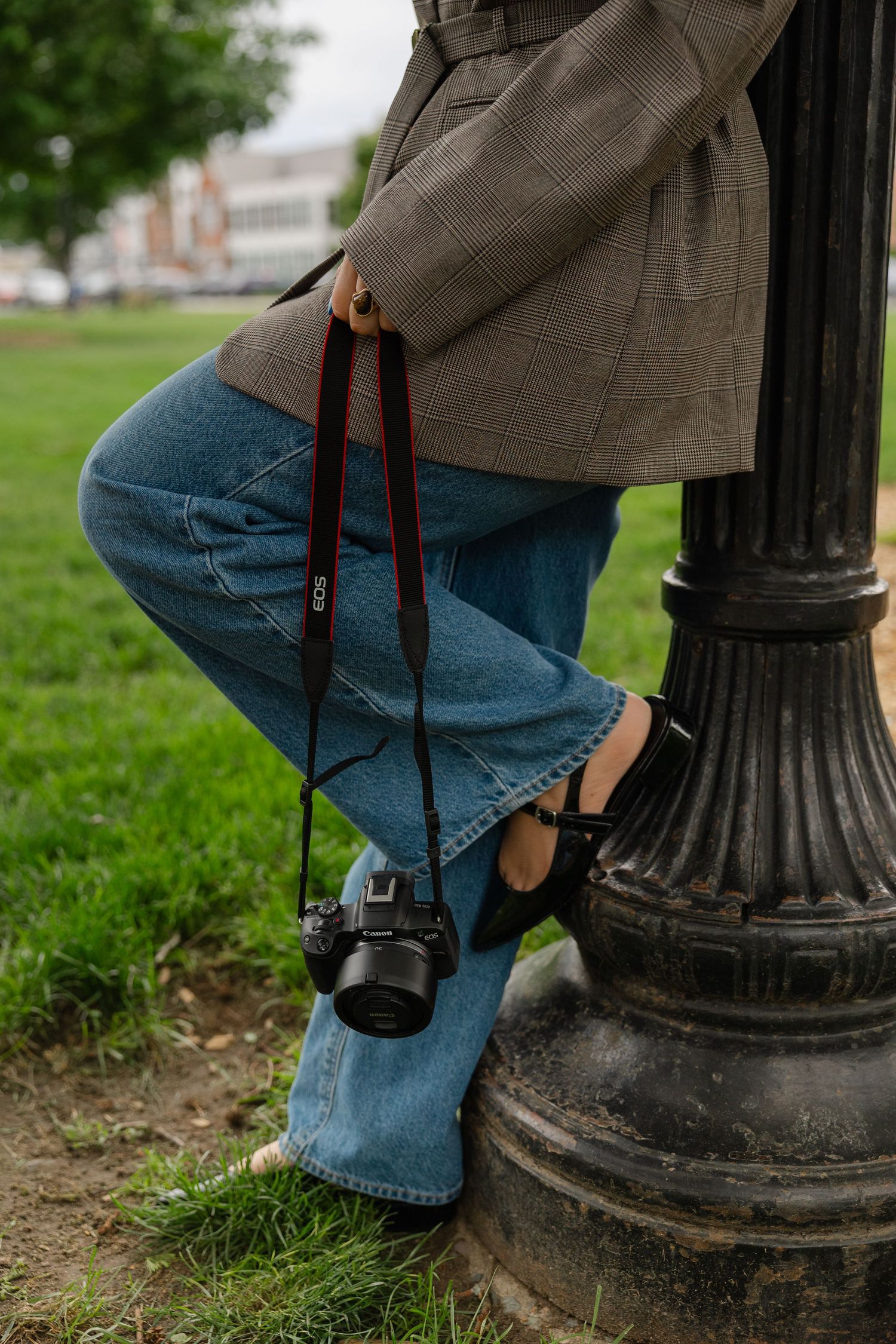 Close-up of photographer in plaid blazer and jeans holding Canon camera by lamppost.