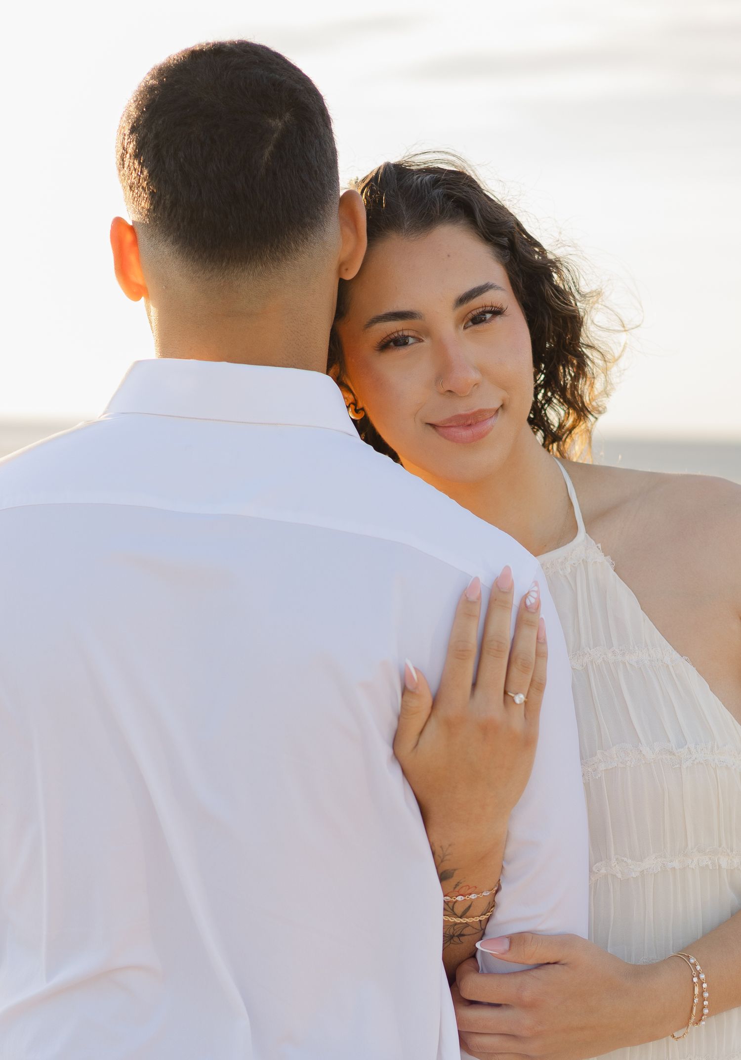 Woman with curly hair in white dress embracing man from behind on beach, showing engagement ring.