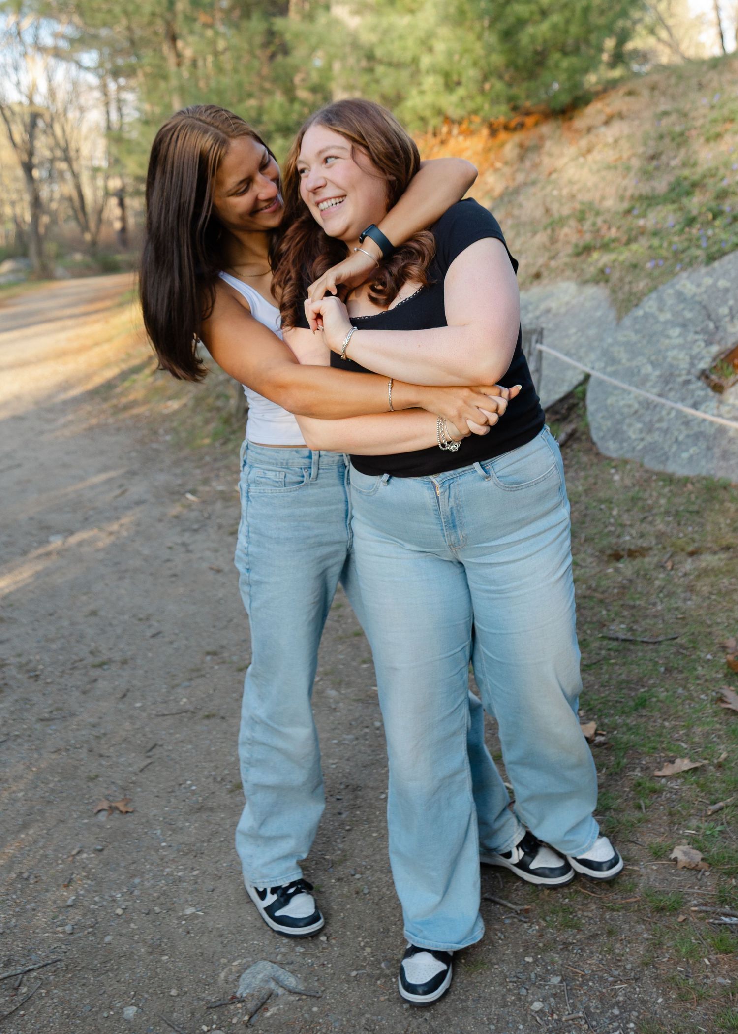 Two young women hugging and laughing together on a nature trail, both wearing light blue jeans and sneakers.