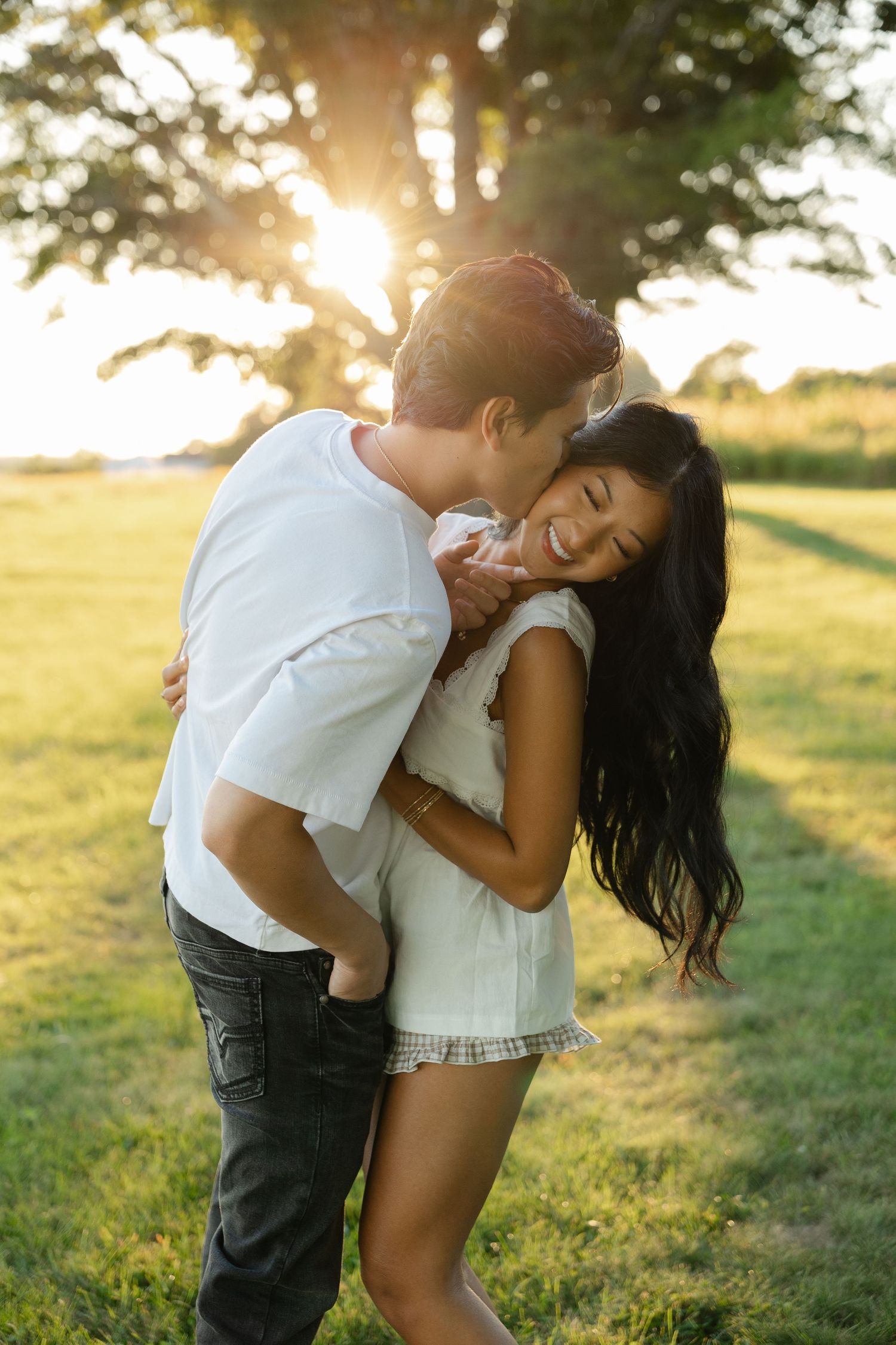 Couple embracing at golden hour in a park, man kissing woman's cheek as she laughs joyfully.