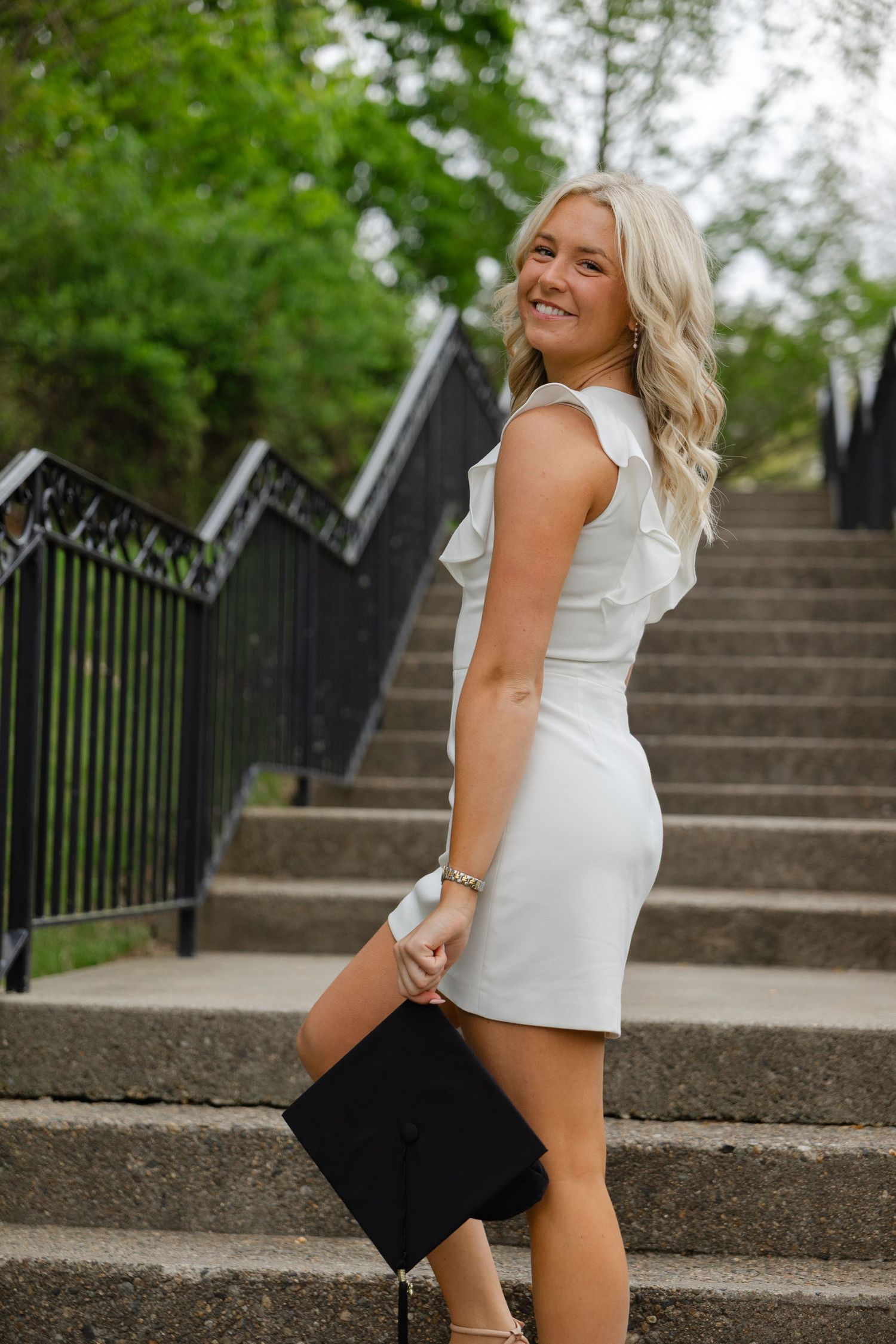 Smiling blonde woman in white ruffle mini dress holding graduation cap on outdoor stone steps.