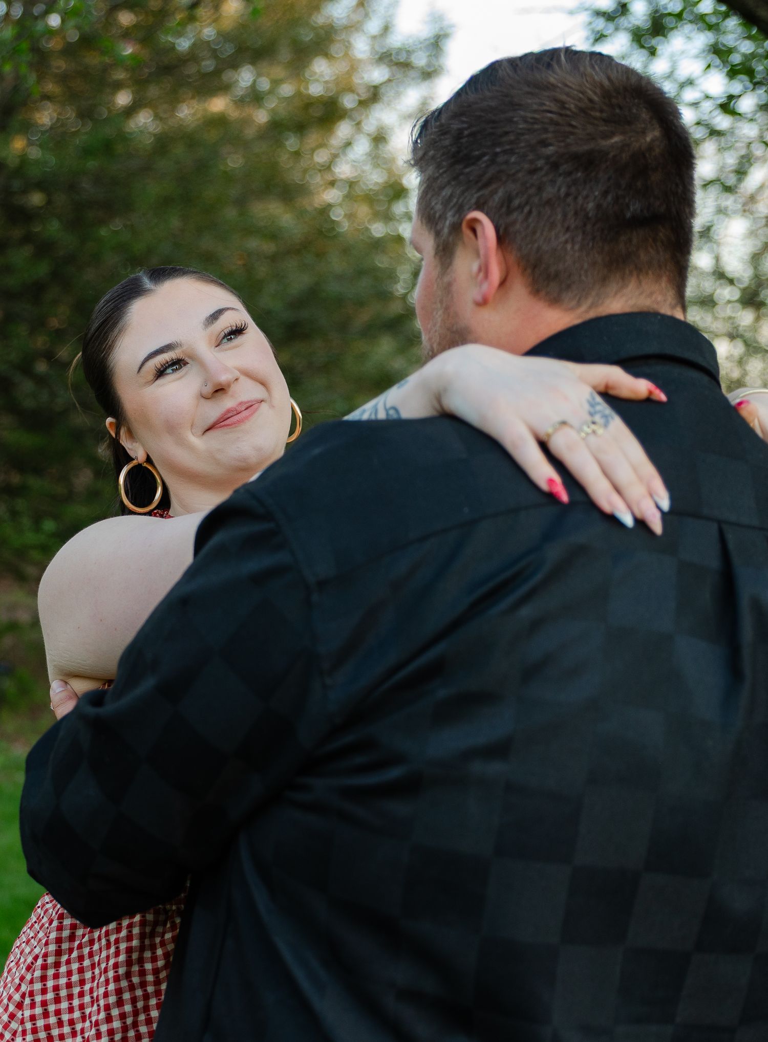 A couple embraces outdoors, woman in red gingham smiles over man's shoulder wearing a black checkered shirt.