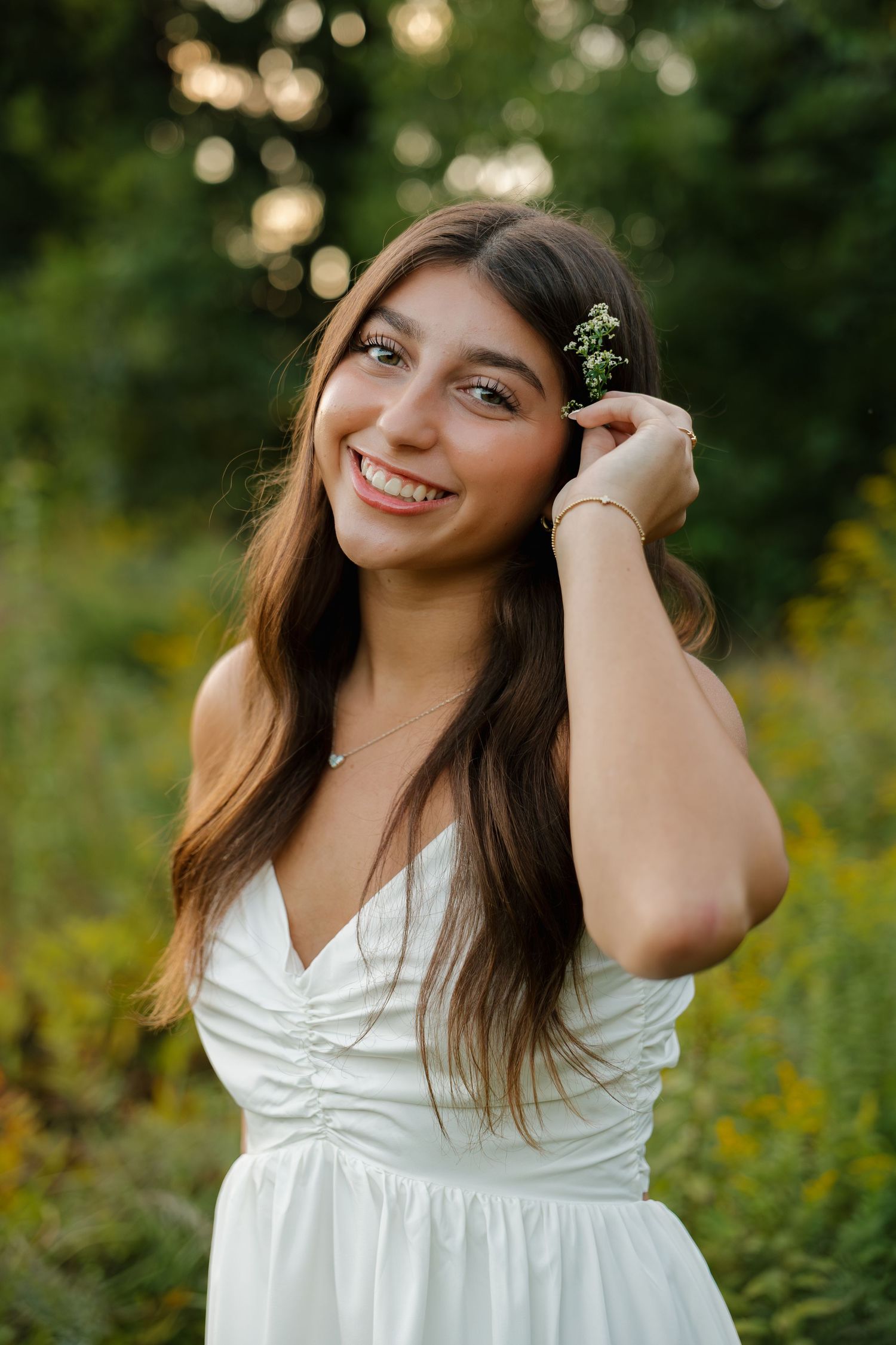 Smiling young woman in white dress holding small wildflowers near her ear in a sunlit green outdoor setting.