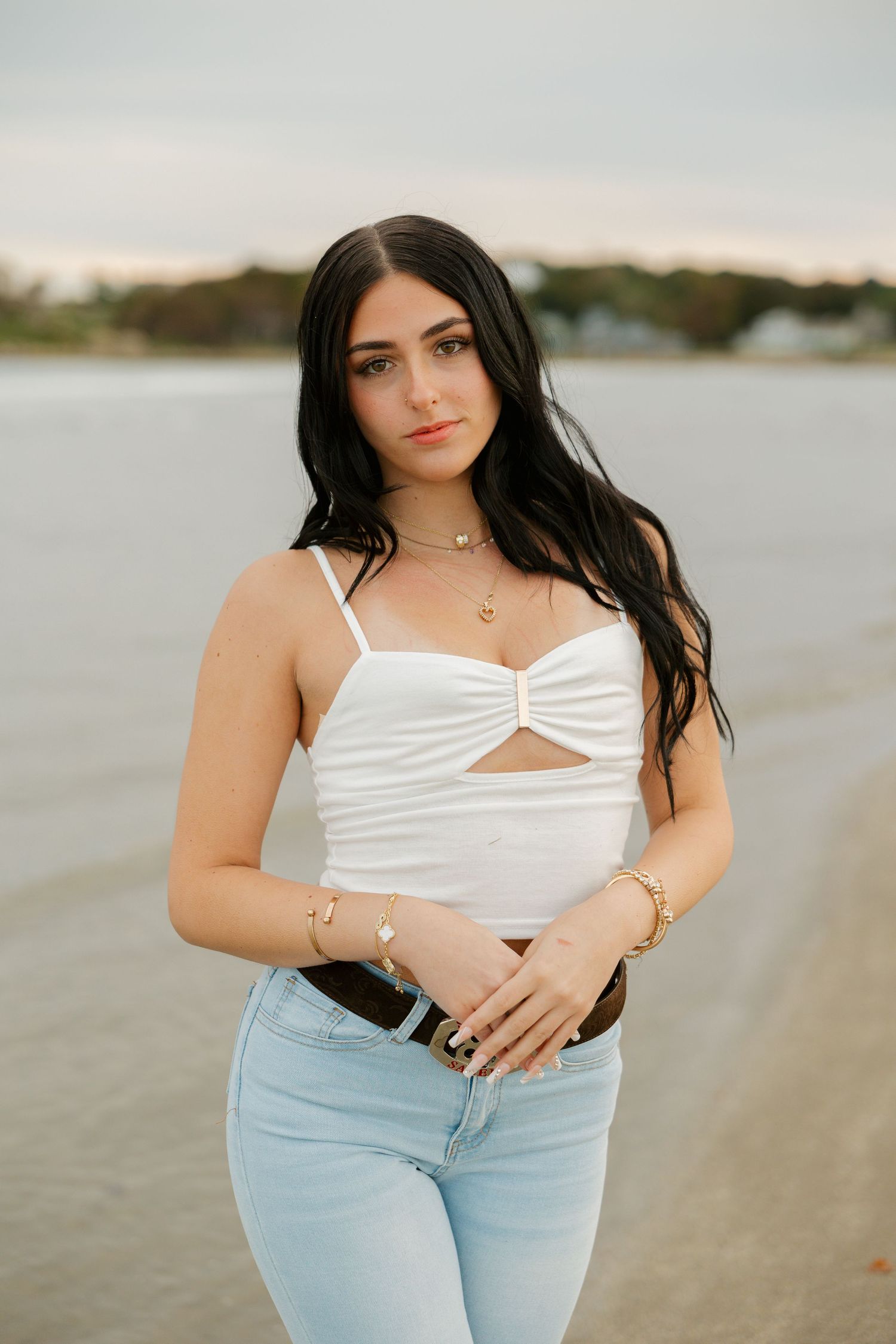 Young woman in white crop top and light jeans posing on a beach at sunset with long dark hair.
