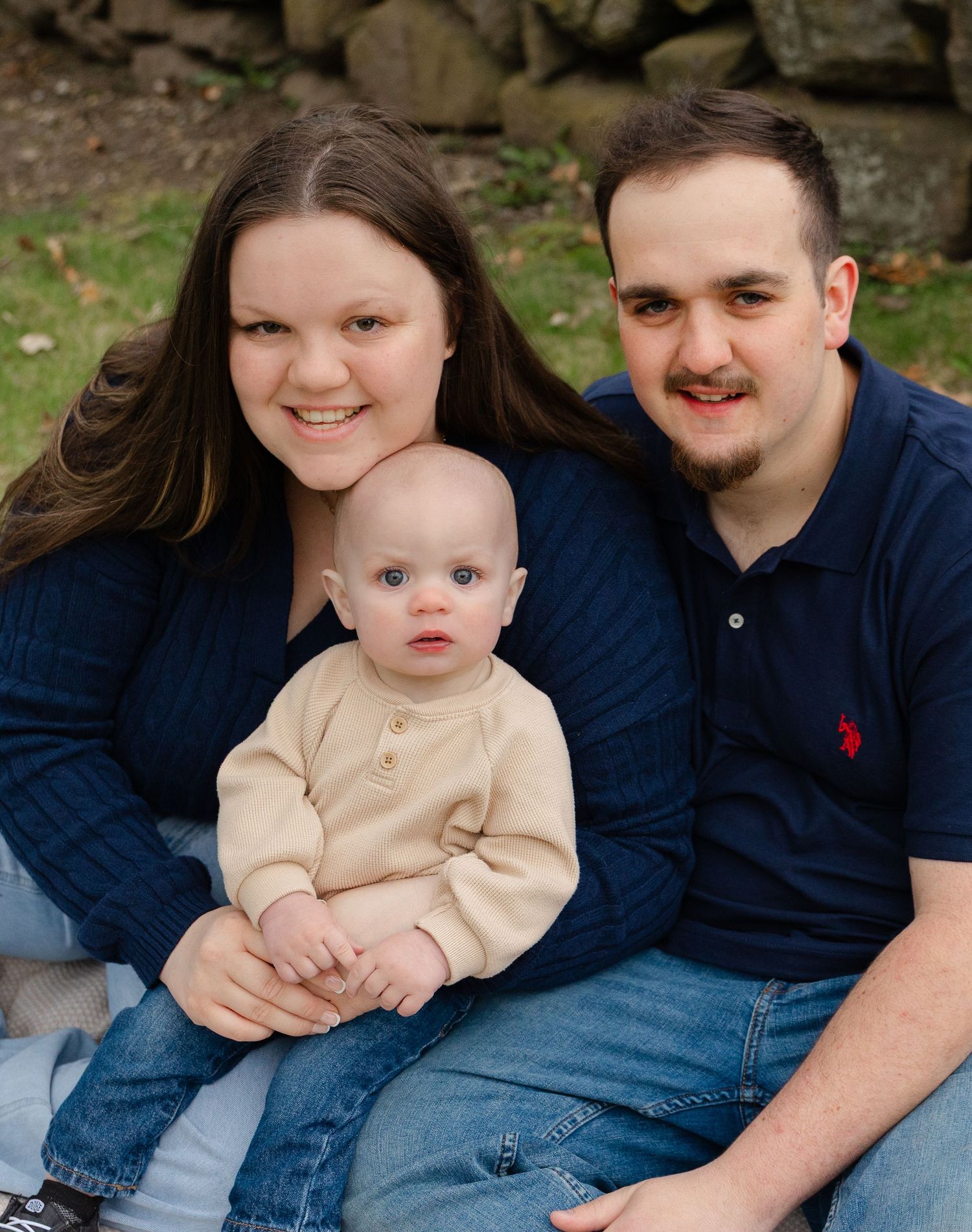 A young family poses outdoors, with a smiling couple in navy blue holding an alert baby in a cream outfit.