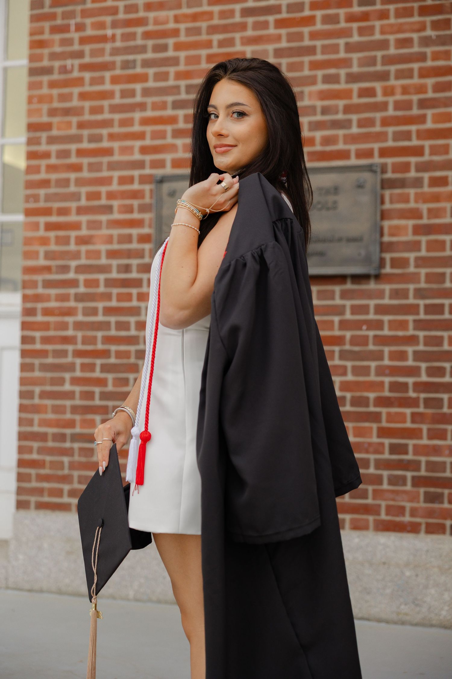 Young woman in black graduation gown and cap smiling outdoors near brick building.