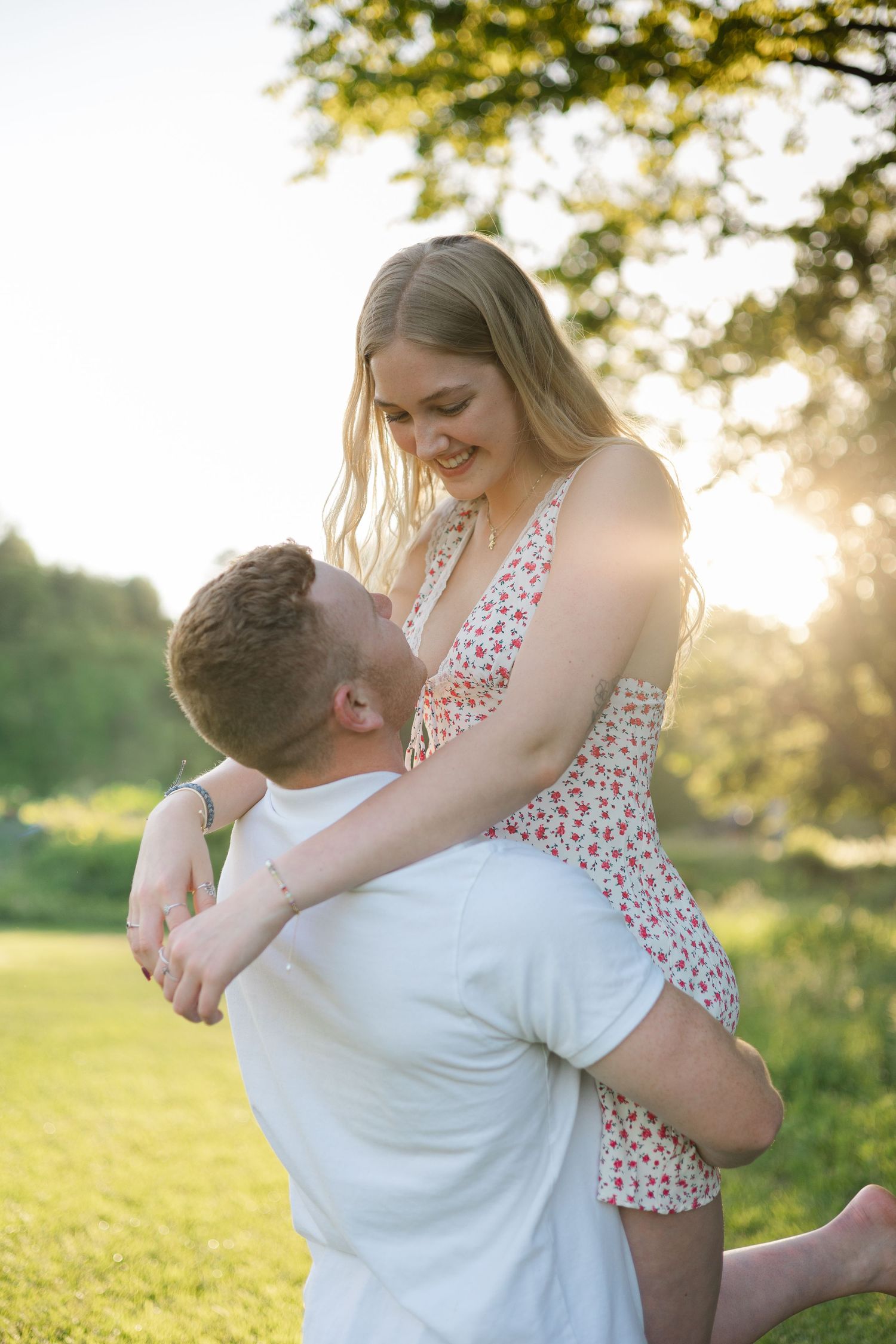 A couple shares a romantic moment outdoors, man lifting woman in floral dress, bathed in golden sunset light.