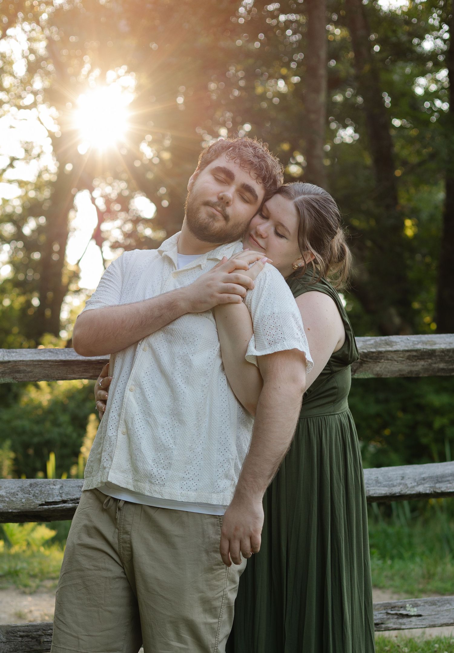 Couple embracing at golden hour near a rustic wooden fence surrounded by lush green trees in a romantic outdoor setting.
