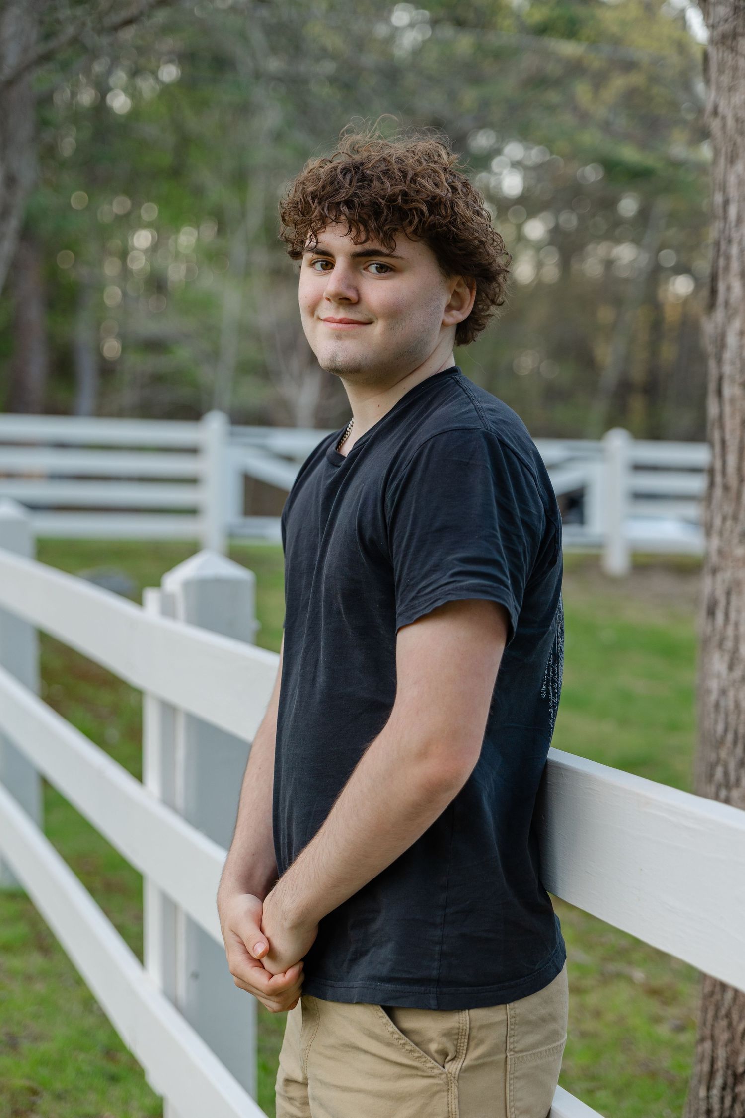 A teenage boy with curly hair smiles while leaning against a white fence outdoors, wearing a black t-shirt and khaki shorts.