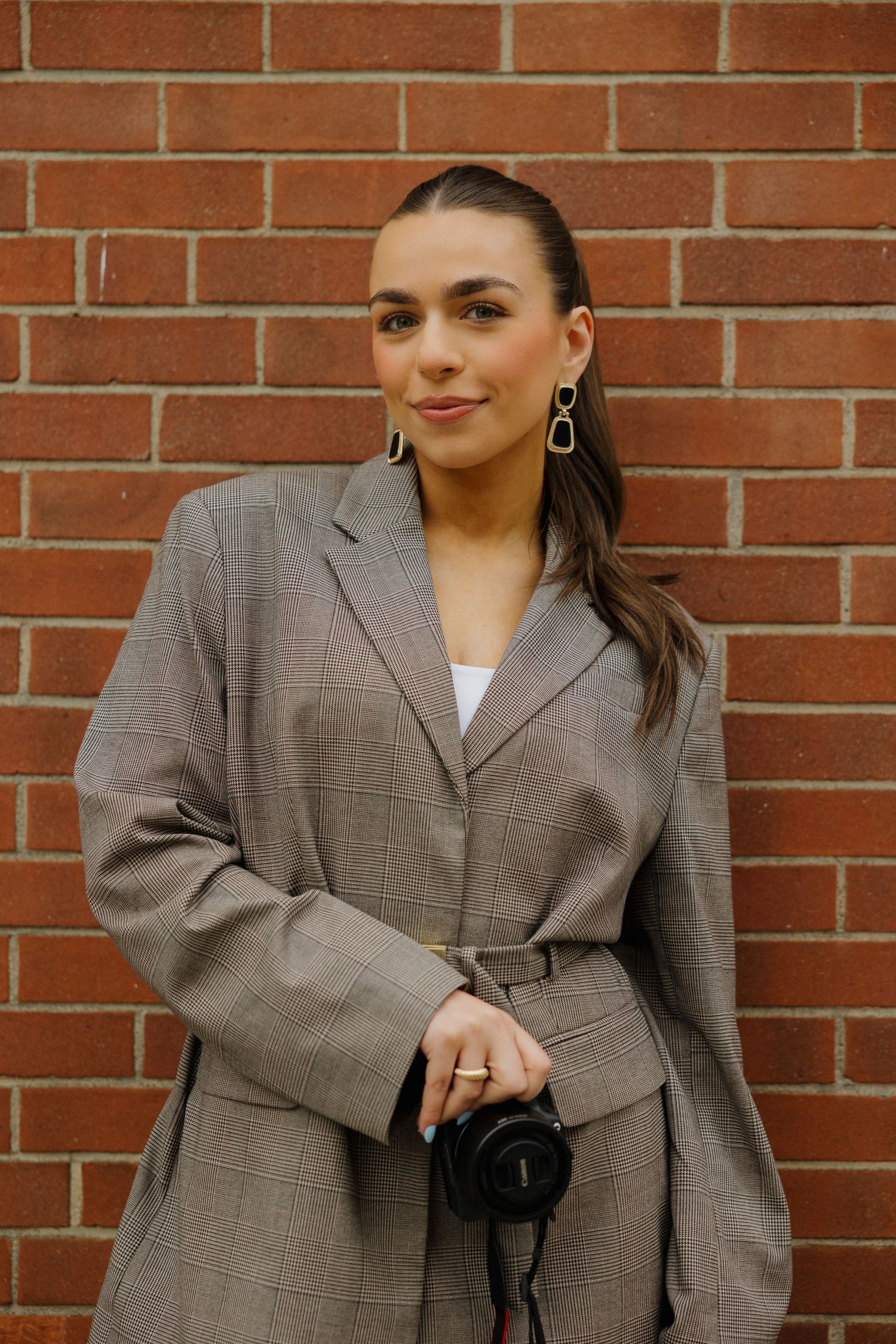 Woman in plaid blazer holding camera against brick wall, wearing gold earrings and ring, smiling confidently.