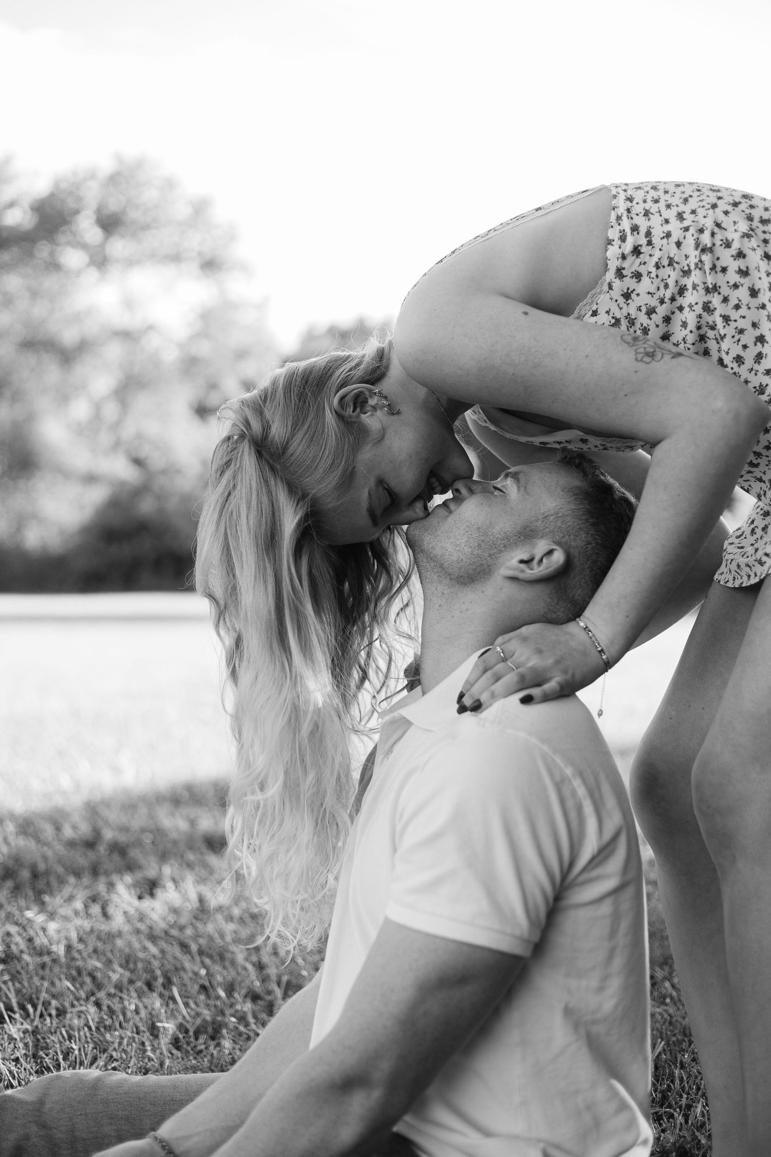 Black and white photo of a romantic couple kissing outdoors, woman leaning down to kiss man sitting on grass.