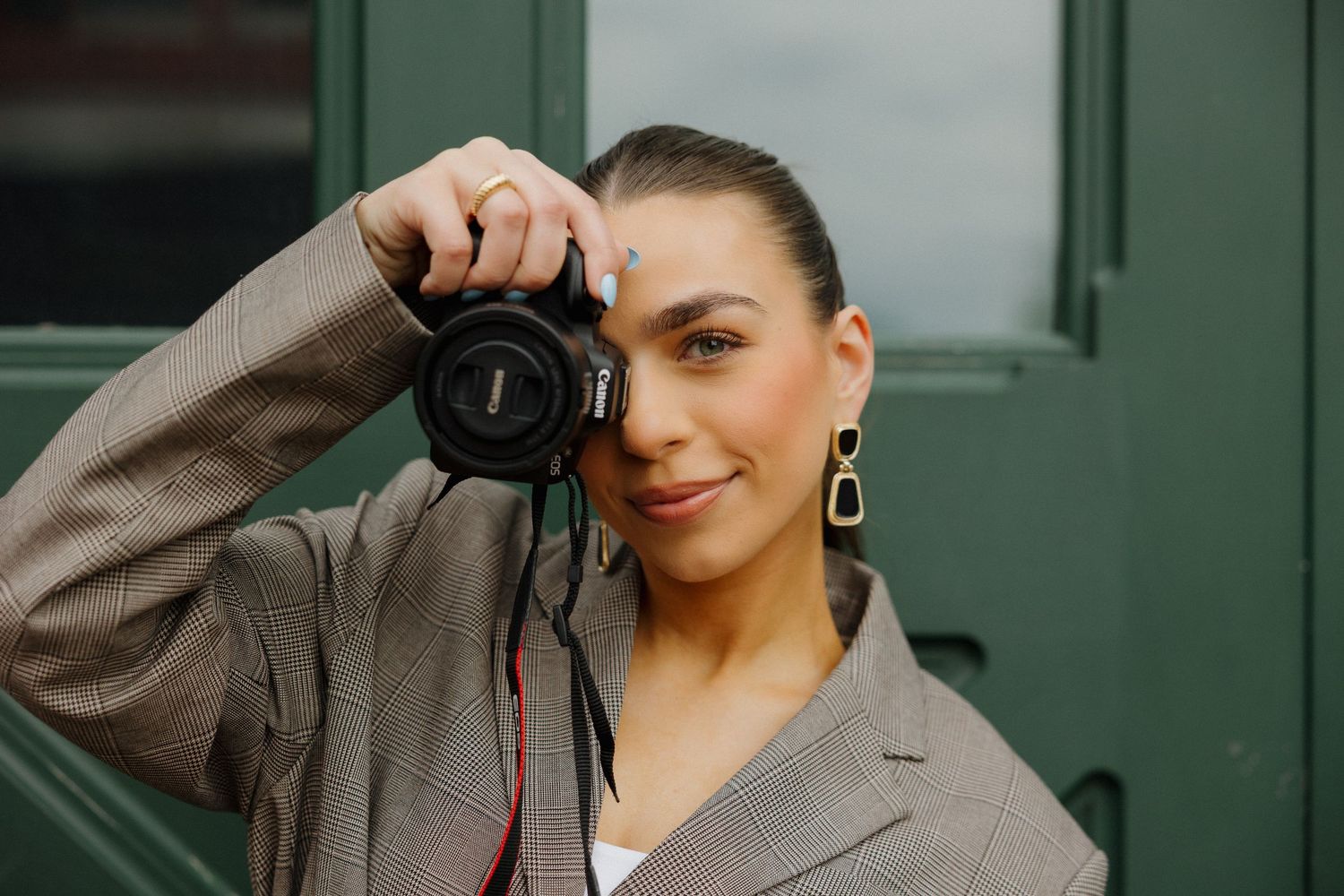 A young female photographer holds a Canon camera up to her eye, wearing a gray blazer and gold earrings.