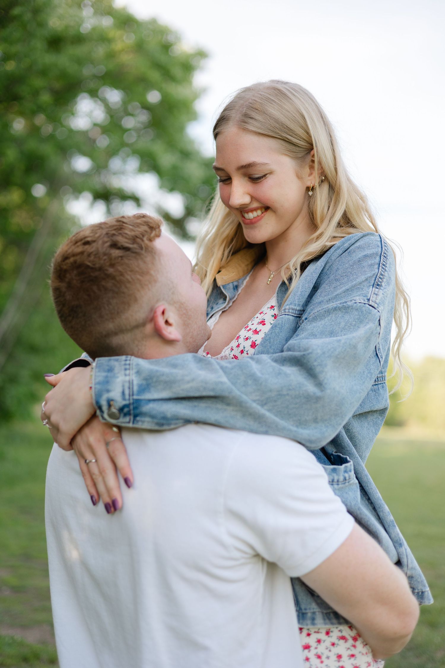 Happy blonde woman in floral dress embracing red-haired man outdoors in sunny green park setting.
