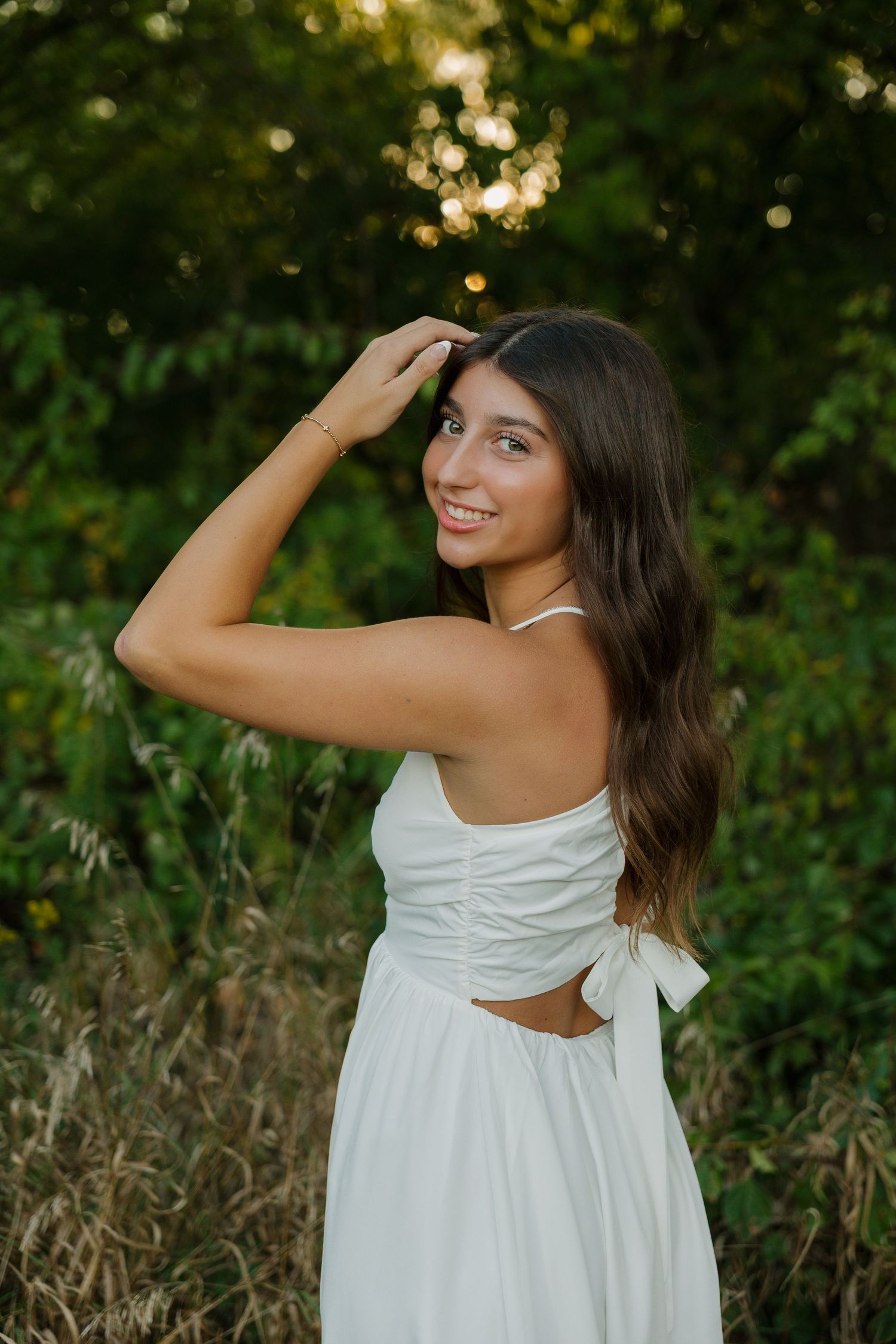 Young woman in white dress smiling outdoors among lush greenery at golden hour.
