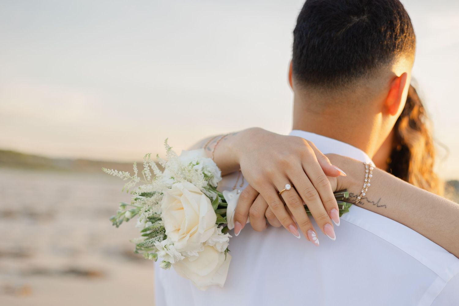 Bride resting hand with engagement ring on grooms shoulder holding white bouquet at beach.