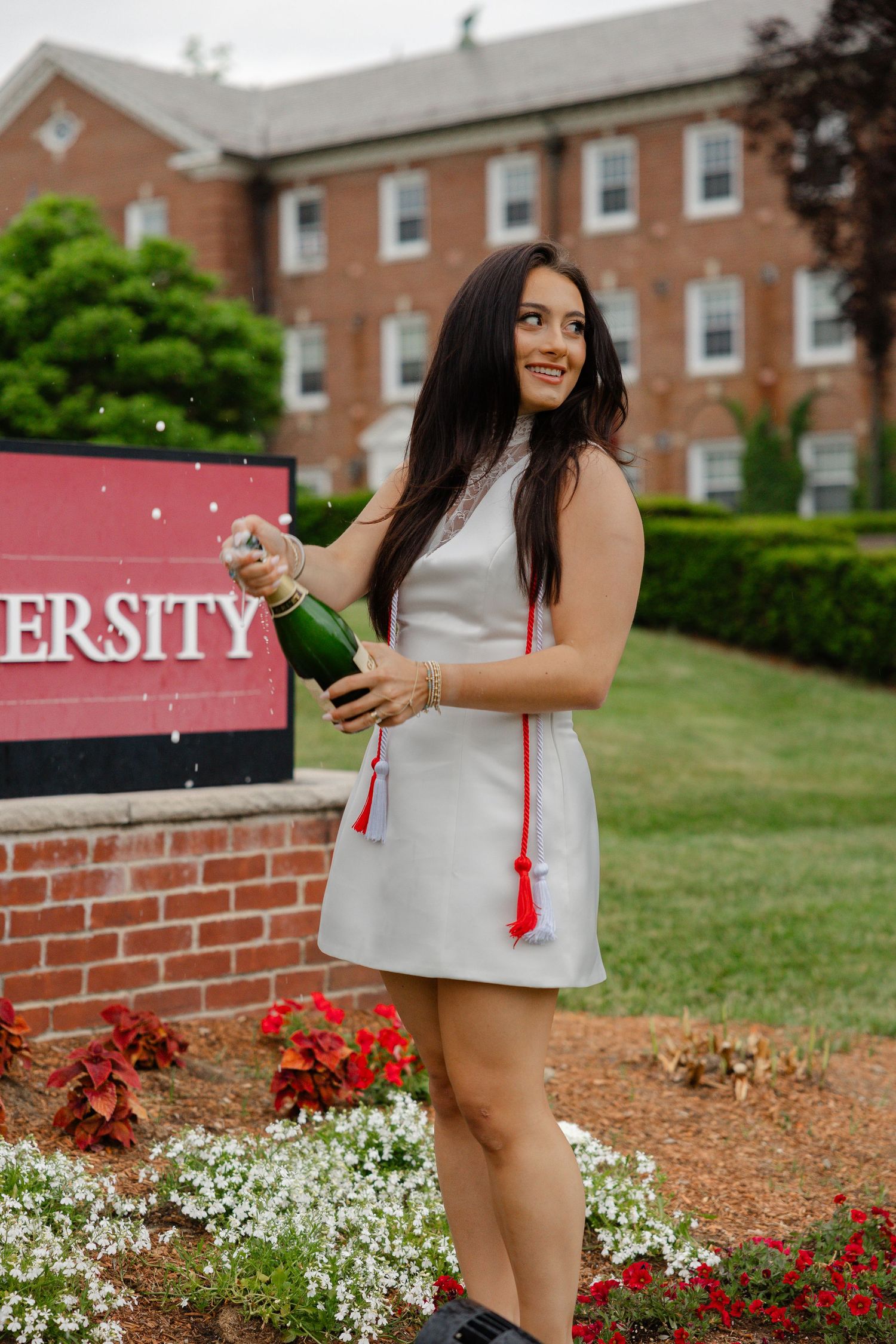 A young woman in a white dress pops champagne by a university sign, celebrating graduation with red tassel.