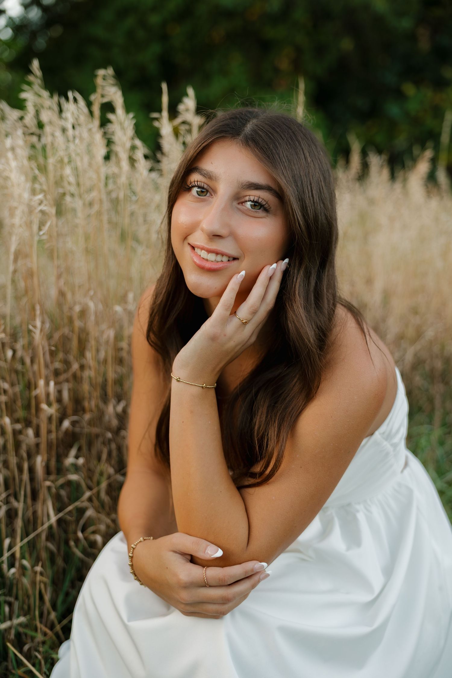 Young woman in white dress sitting in tall golden grass, smiling warmly with hand on cheek, wearing gold jewelry.