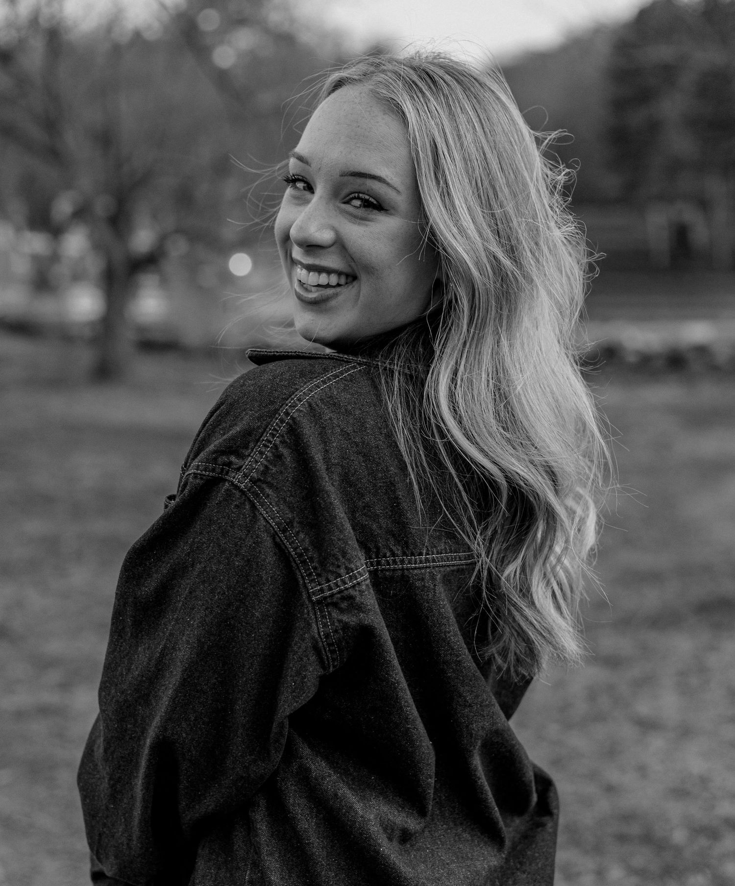 Black and white photo of a smiling blonde woman in a denim jacket turning to look back outdoors.