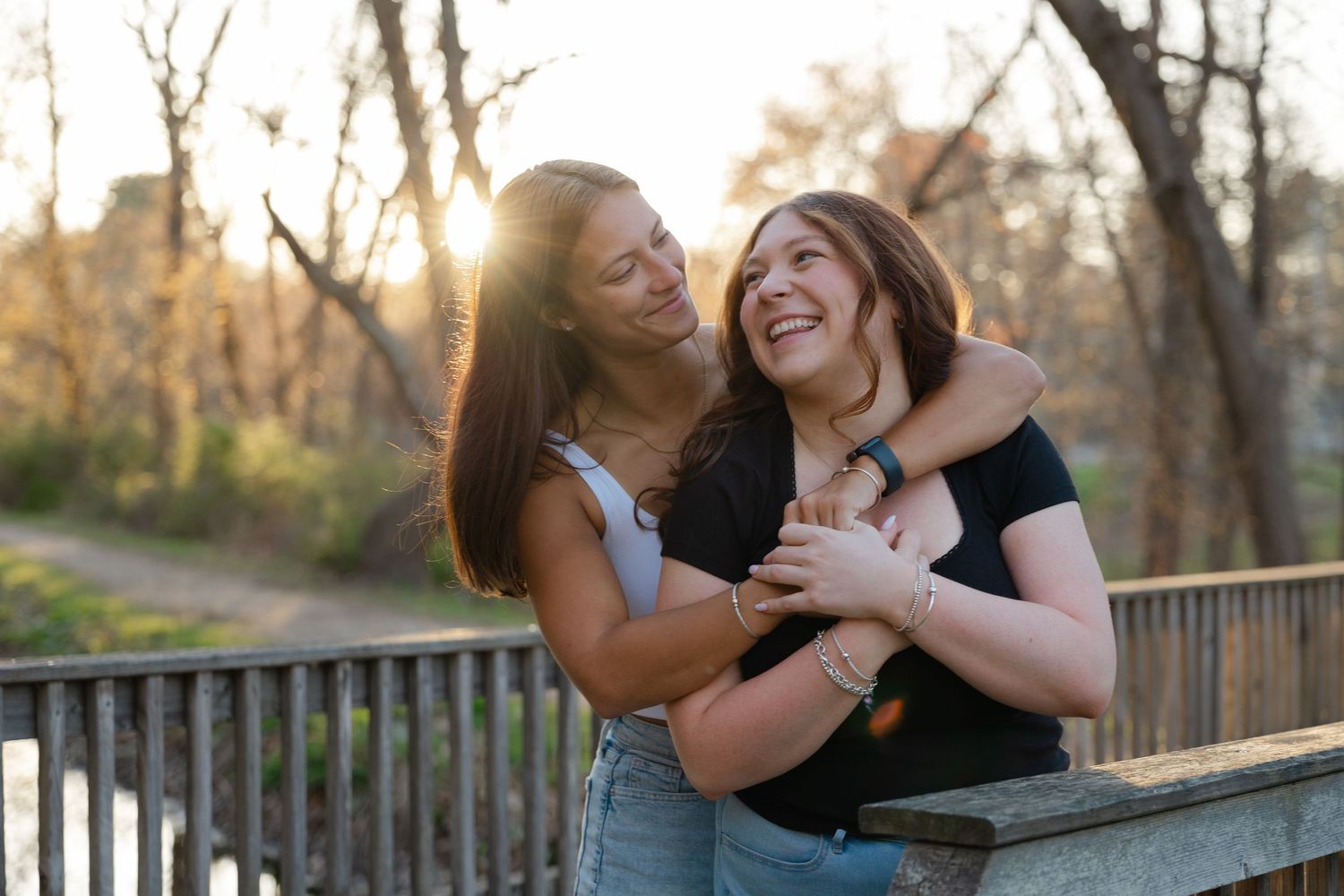 Two young women laughing and embracing on a wooden bridge at golden hour in a wooded park.