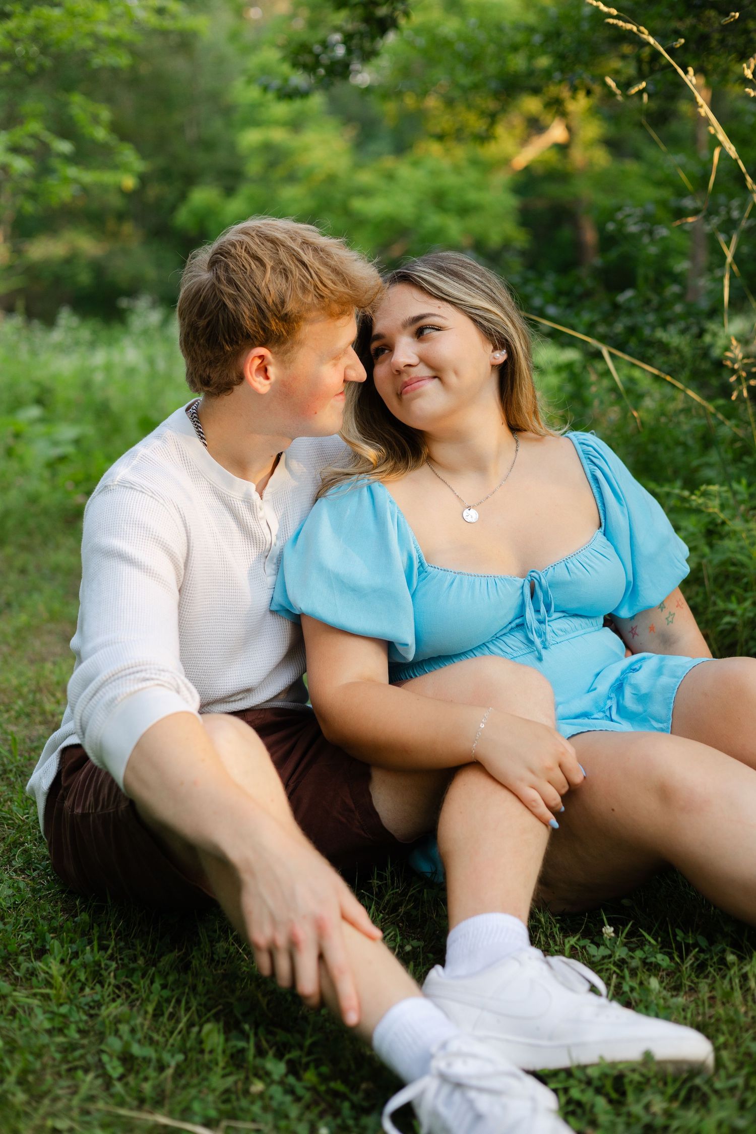 Couple sitting on grass in a park, woman in blue dress smiling at redhead man in white sweater and brown shorts.