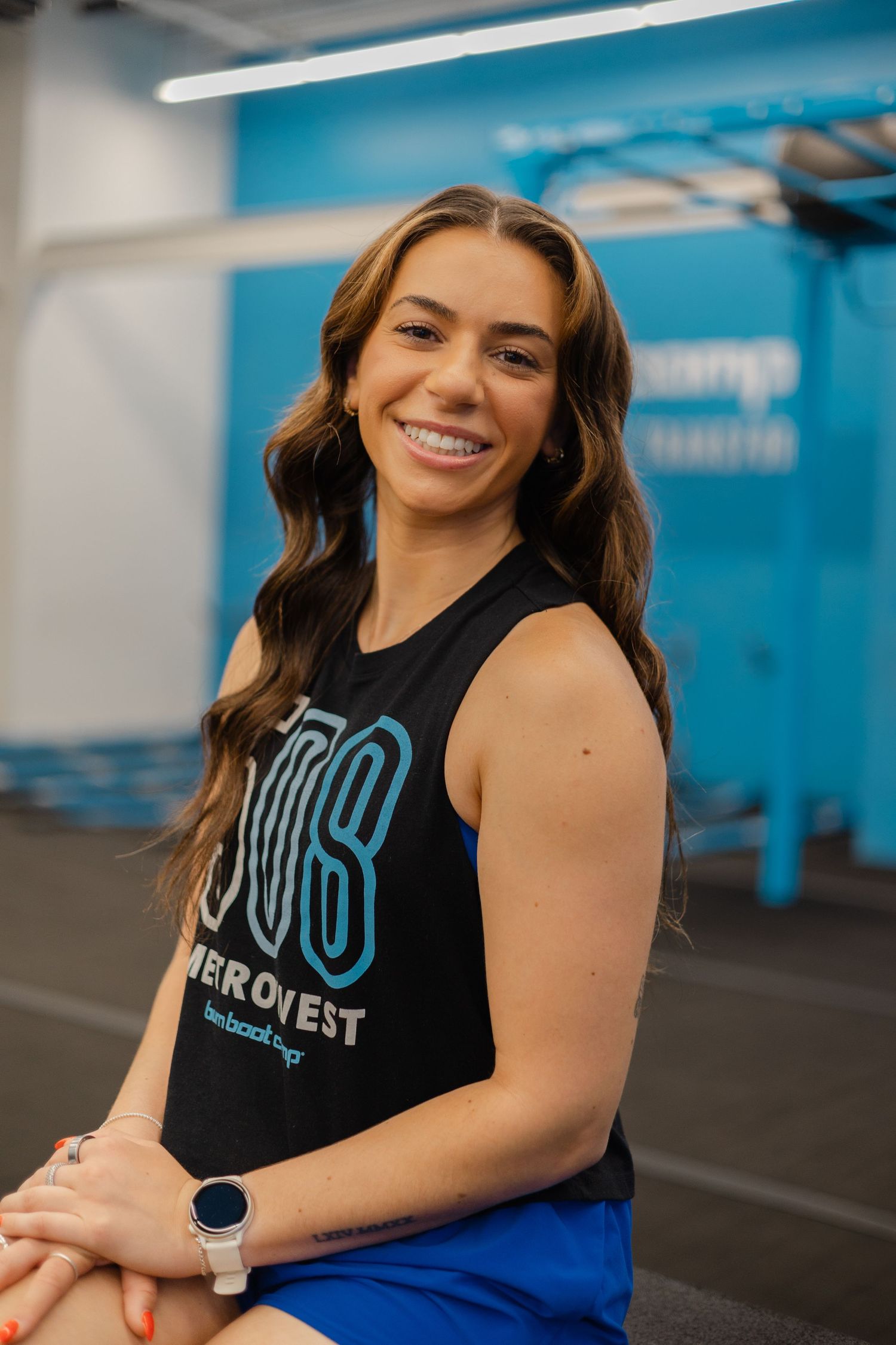 Smiling young woman in a black FreeBootcamp tank top poses in a bright blue gym fitness studio.