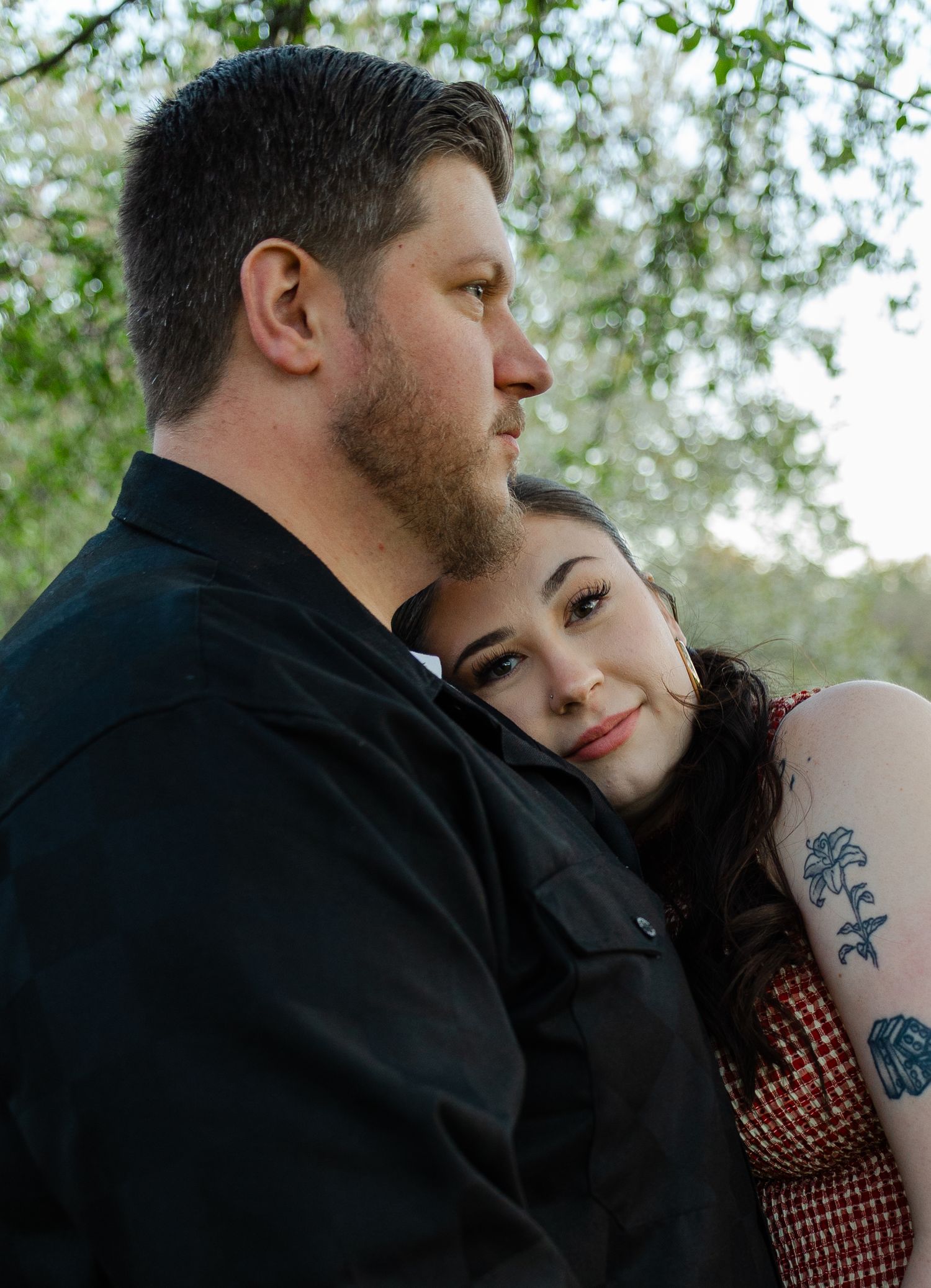 Couple embracing outdoors under green trees, woman with rose tattoo smiling, man in black shirt looking away.