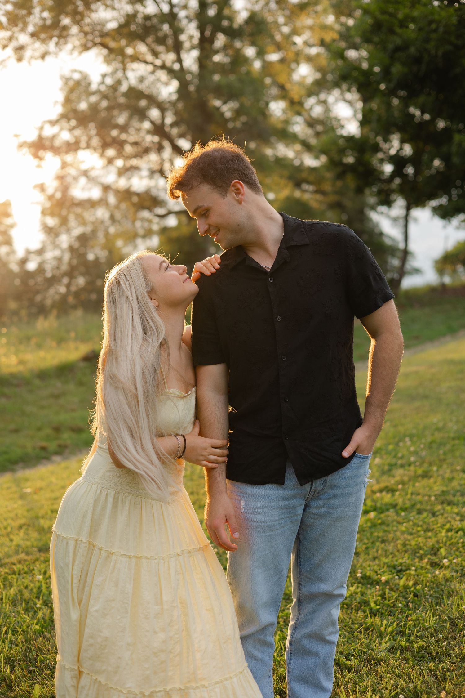 A couple shares a romantic moment at golden hour in a park, woman in yellow dress gazing up at man in black shirt.