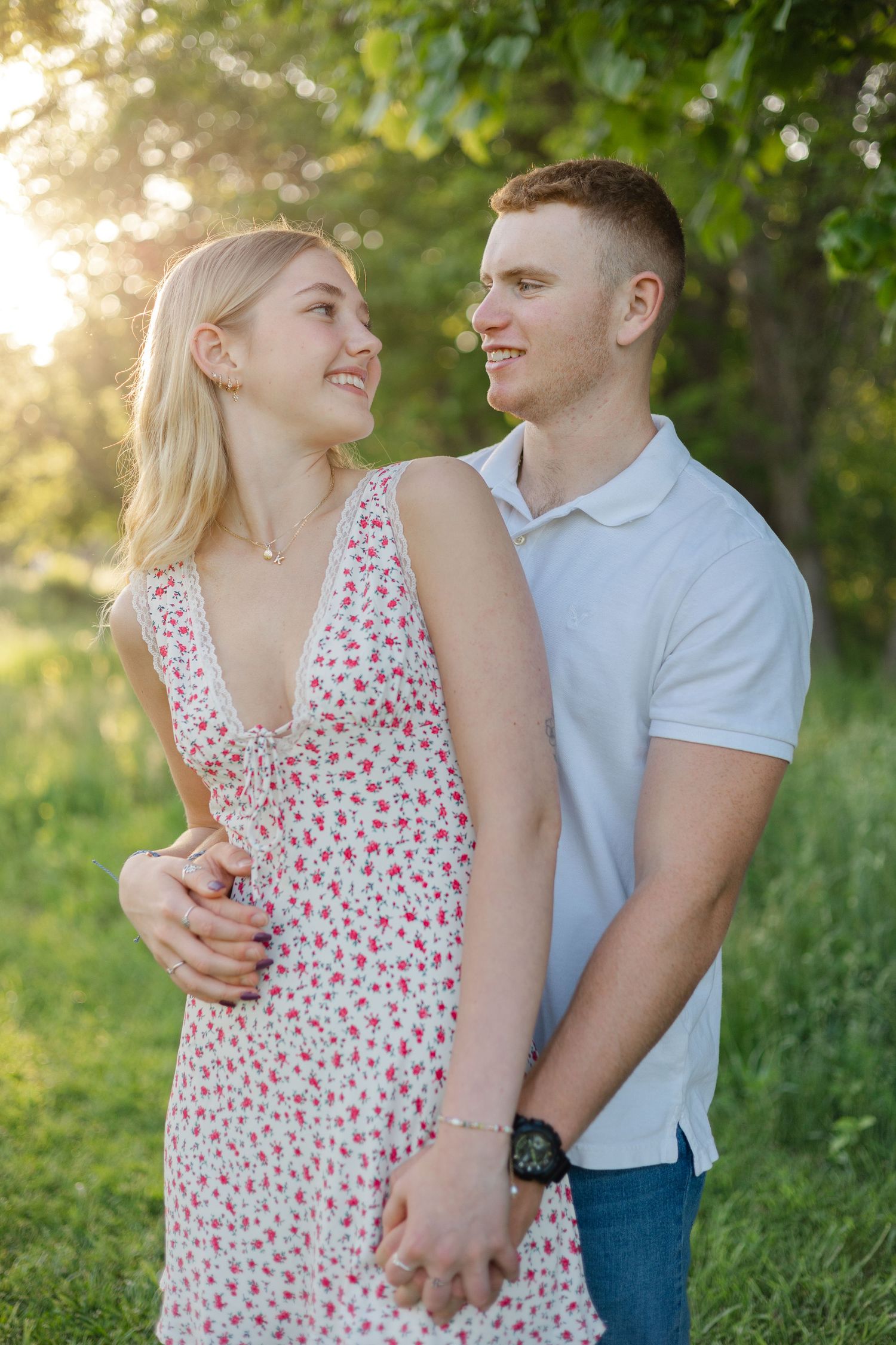 A young couple smiles at each other outdoors at golden hour, she in a floral dress, he in a white polo shirt.
