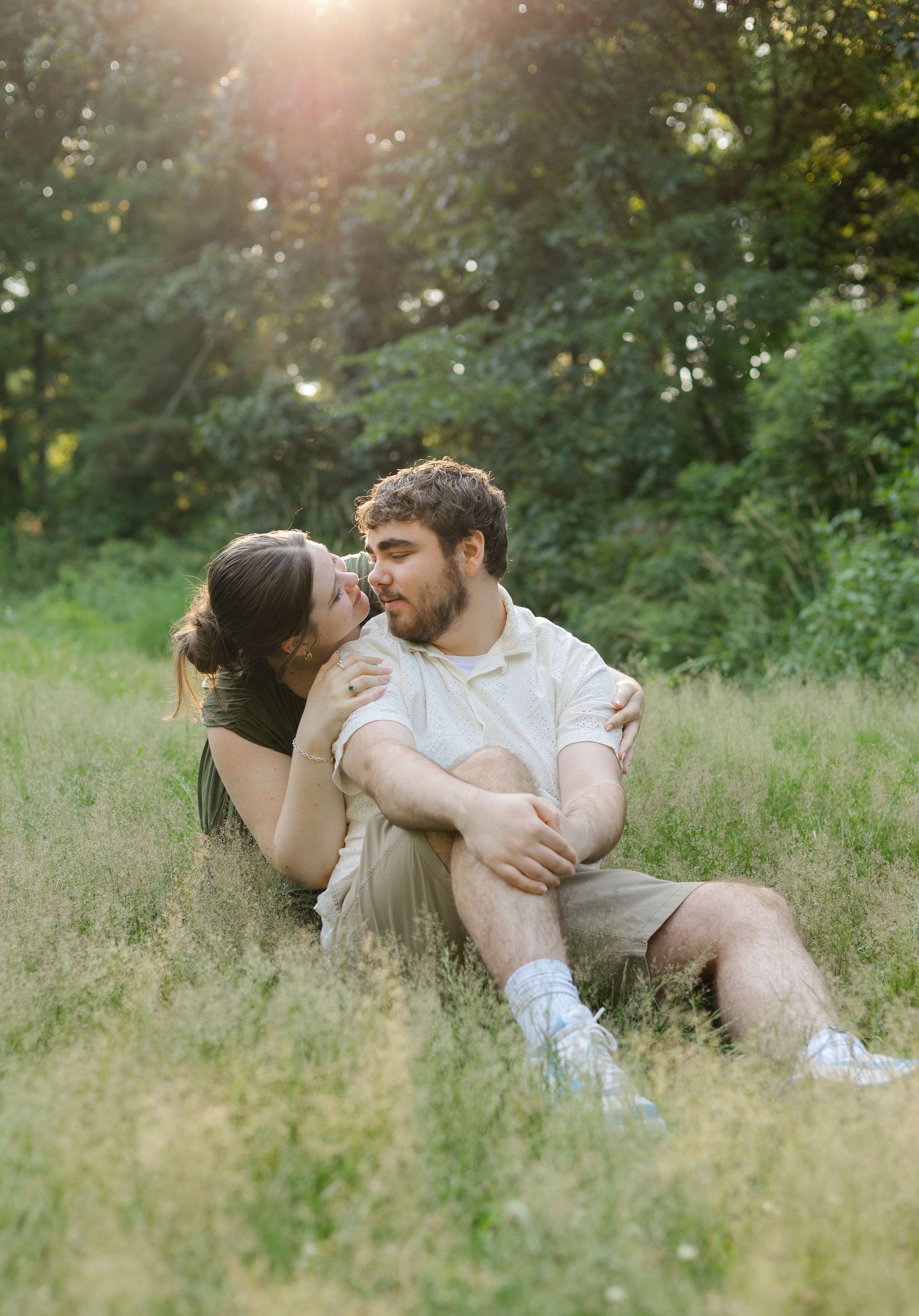 A couple shares a romantic moment sitting in a sunlit grassy meadow surrounded by lush green trees.