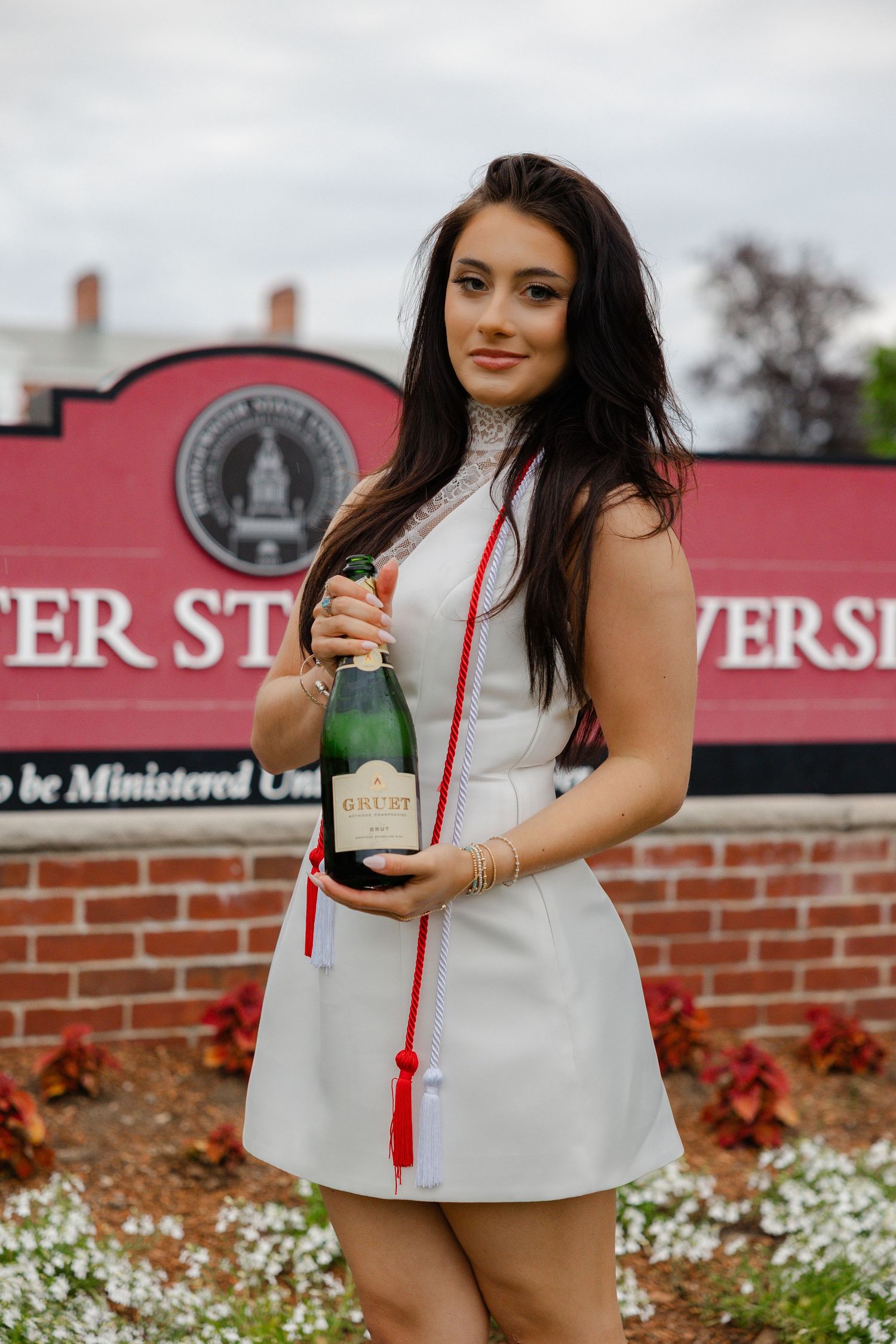 Smiling graduate in white dress holding champagne bottle in front of university sign with red and white graduation stole.