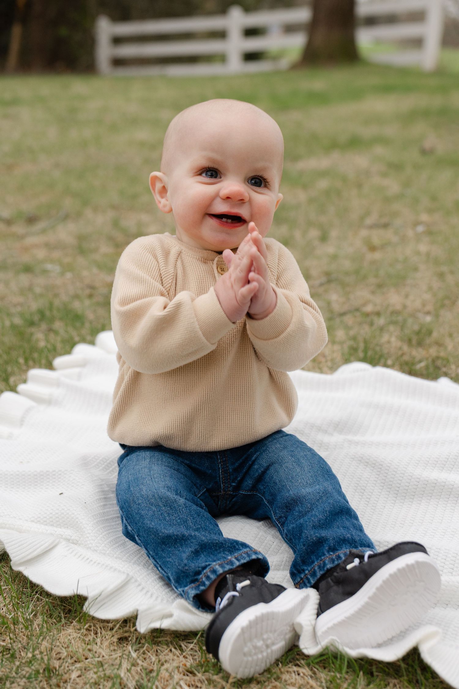 Smiling baby in beige sweater and jeans sits on white blanket outdoors, clapping hands together.