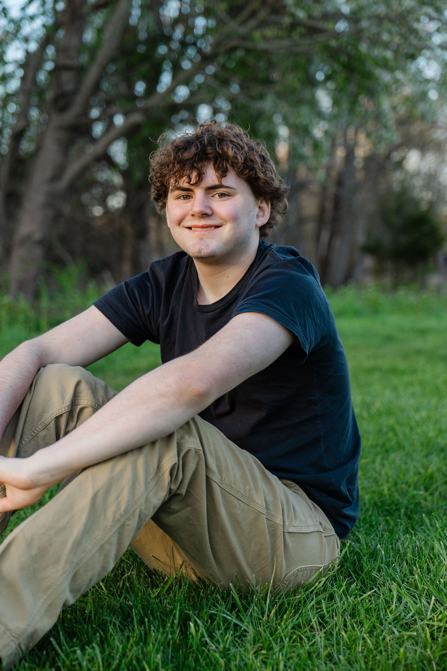 Teenage boy with curly hair sitting cross-legged on green grass, smiling, wearing black t-shirt and khaki pants outdoors.