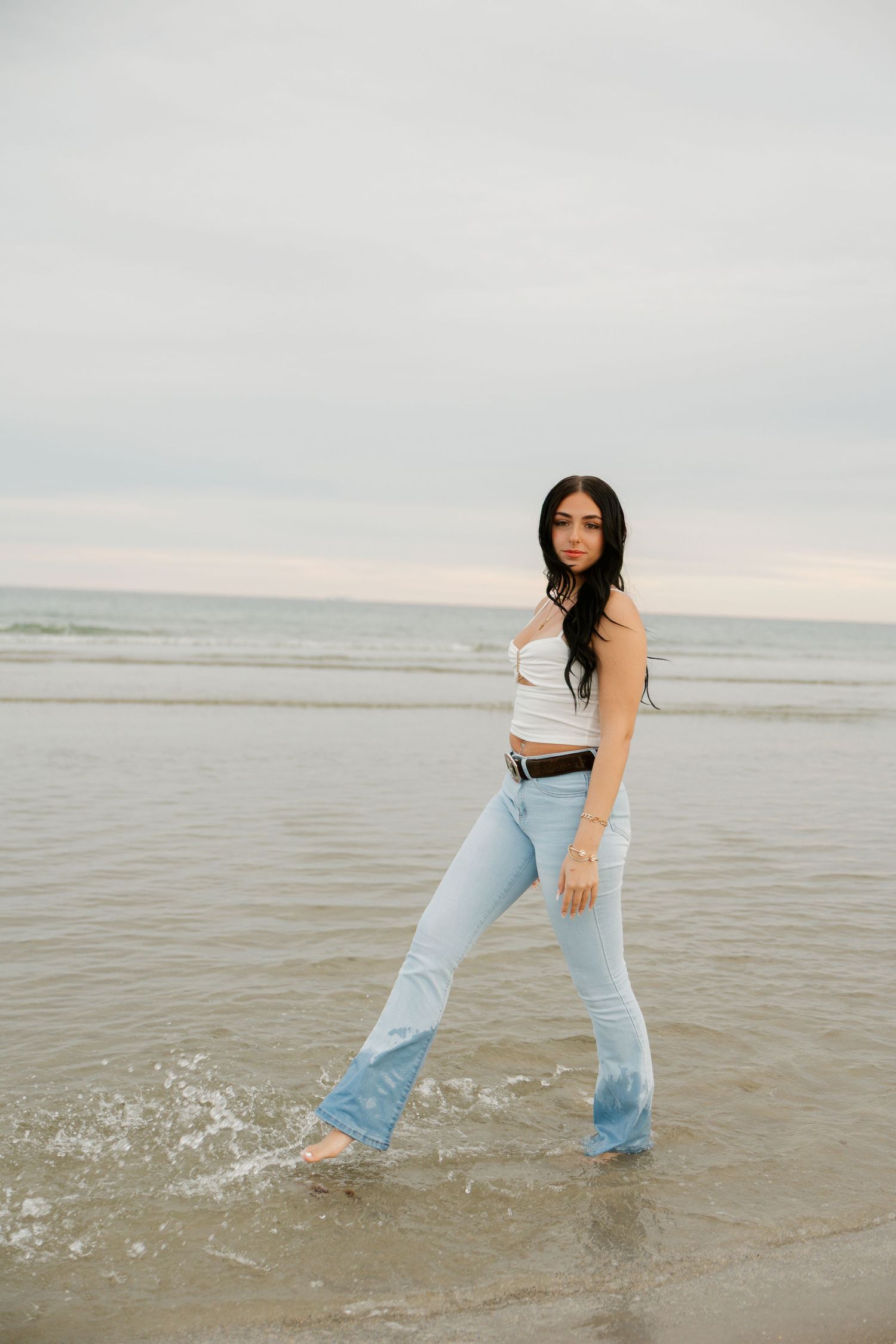 Young woman in white top and light blue flare jeans wading barefoot in ocean waves on a cloudy beach day.