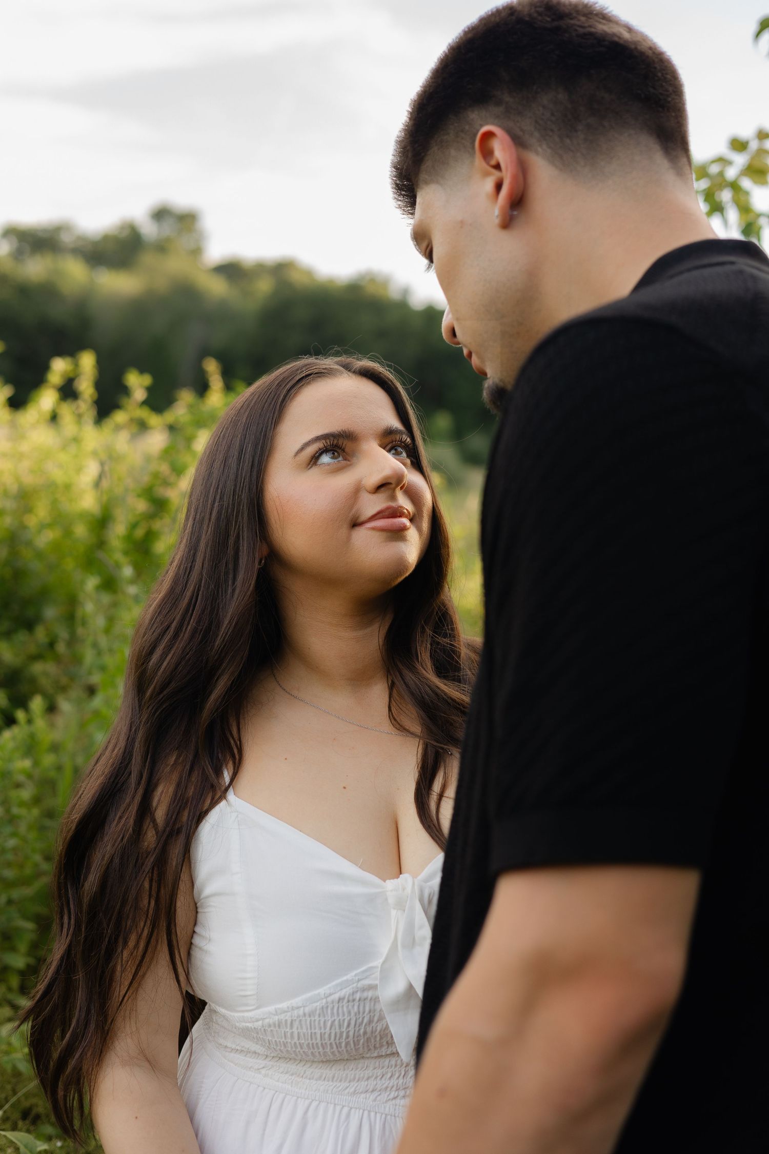 A young couple gazes at each other in a lush green field, woman in white dress smiling up at man in black shirt.