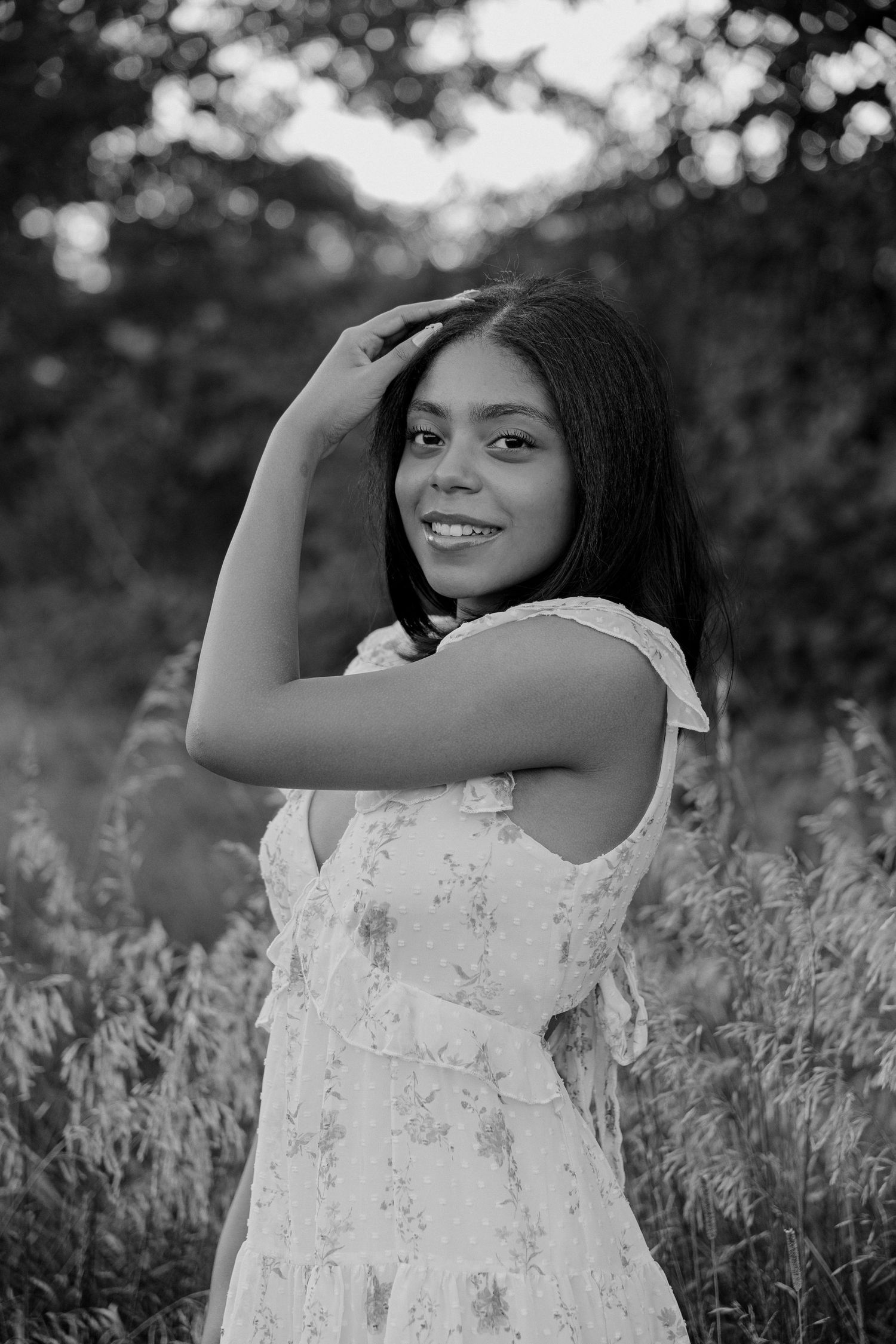 Black and white portrait of a young woman in a floral dress smiling outdoors in a field.