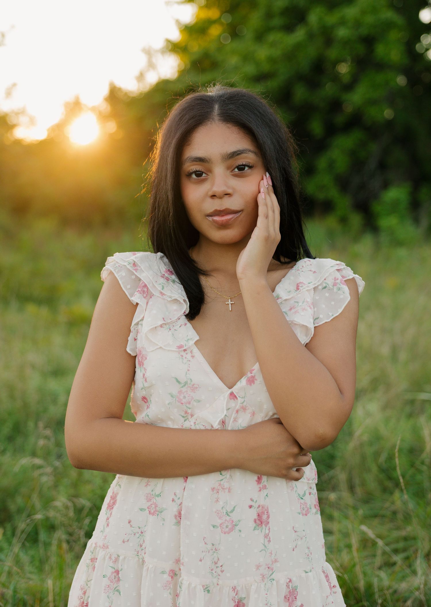 Young woman in floral dress poses at golden hour in a lush green field, wearing a cross necklace.