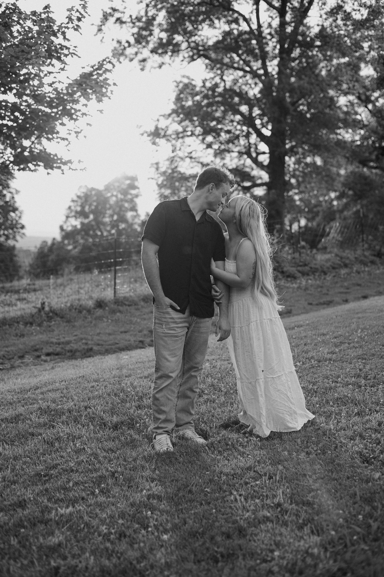 Black and white photo of a couple sharing a romantic kiss outdoors on a grassy path surrounded by trees.