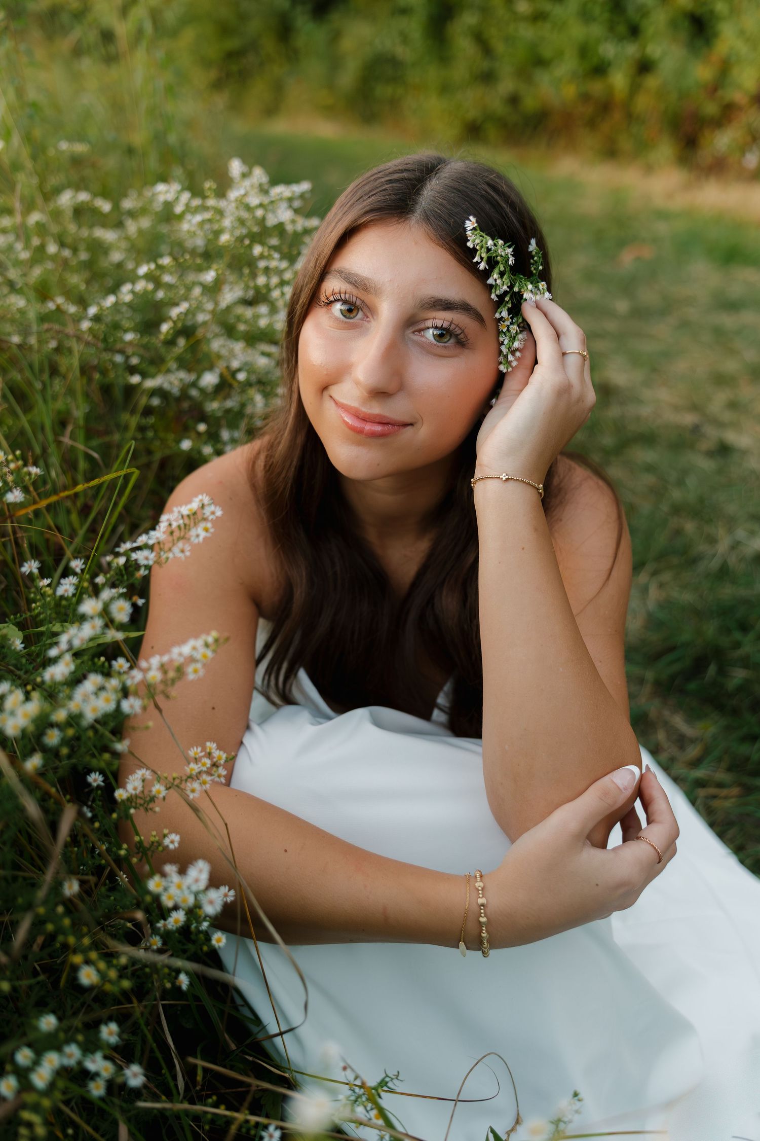 Young woman in white dress holding small white wildflowers in her hair, sitting in a lush green meadow.