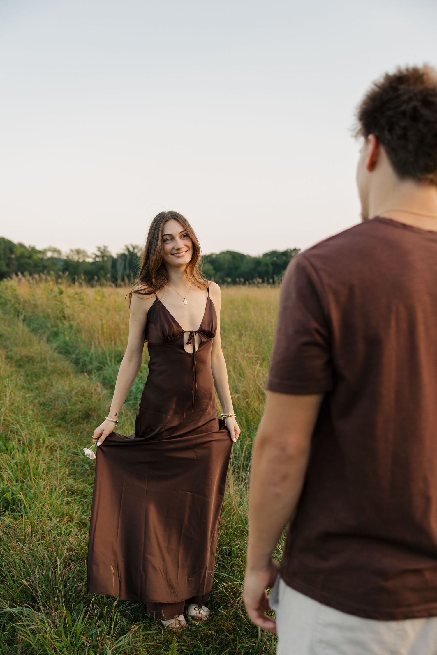 A smiling woman in a brown satin dress stands in a green field, facing a man in a matching brown t-shirt.