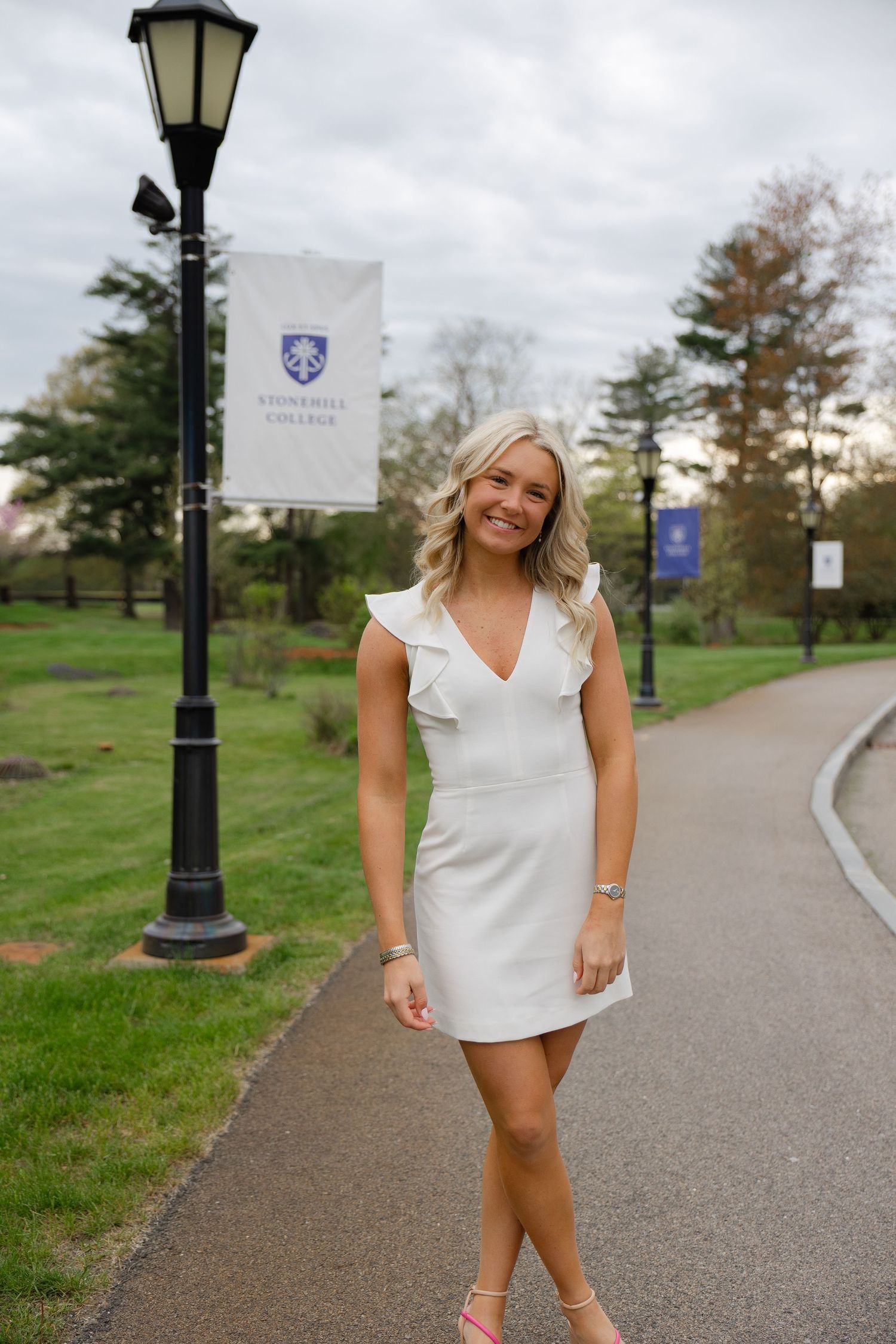 Smiling blonde woman in white ruffle dress poses on a college campus pathway near a lamppost.