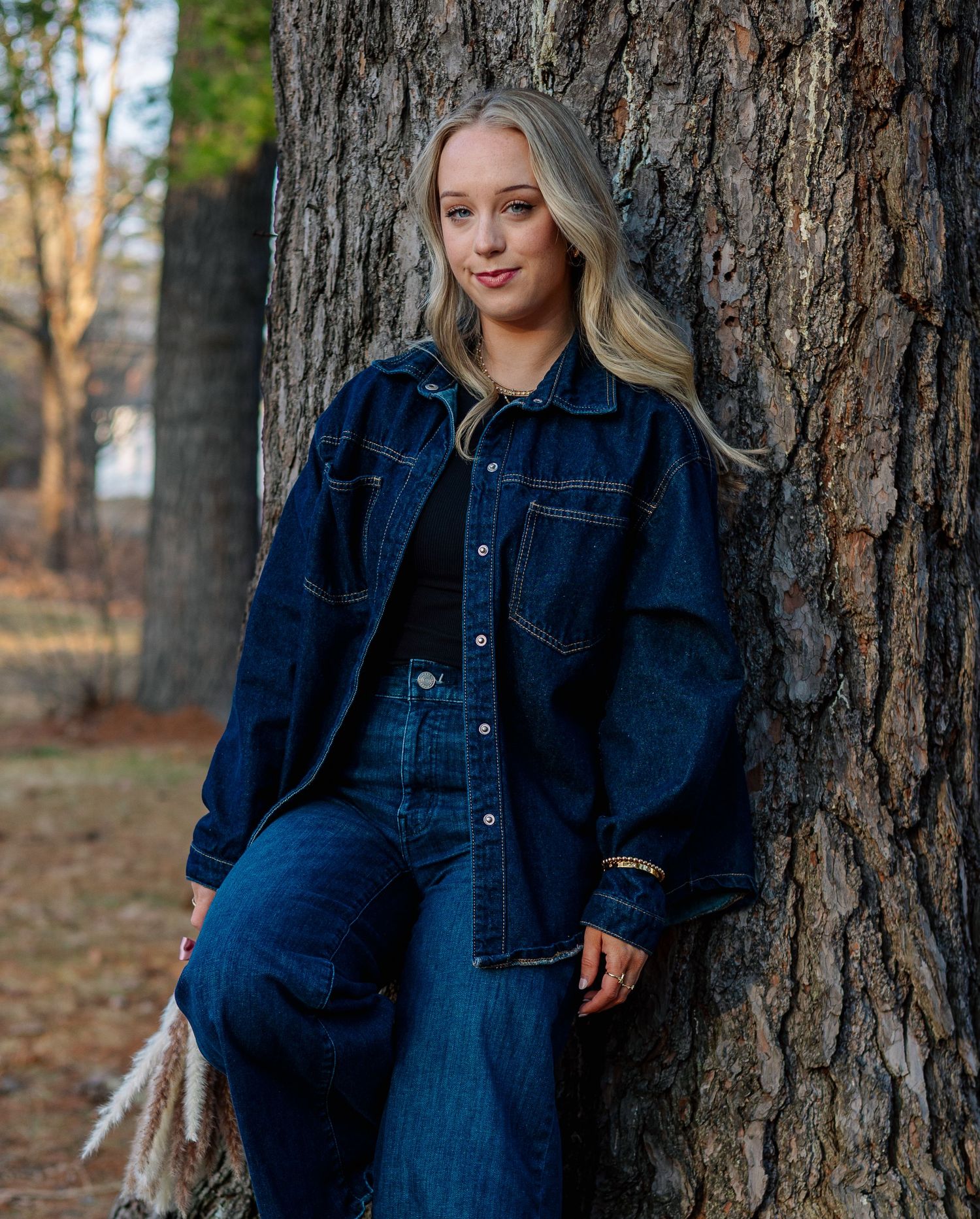 Young blonde woman in dark denim jacket and jeans leaning against a tree trunk outdoors in a park.