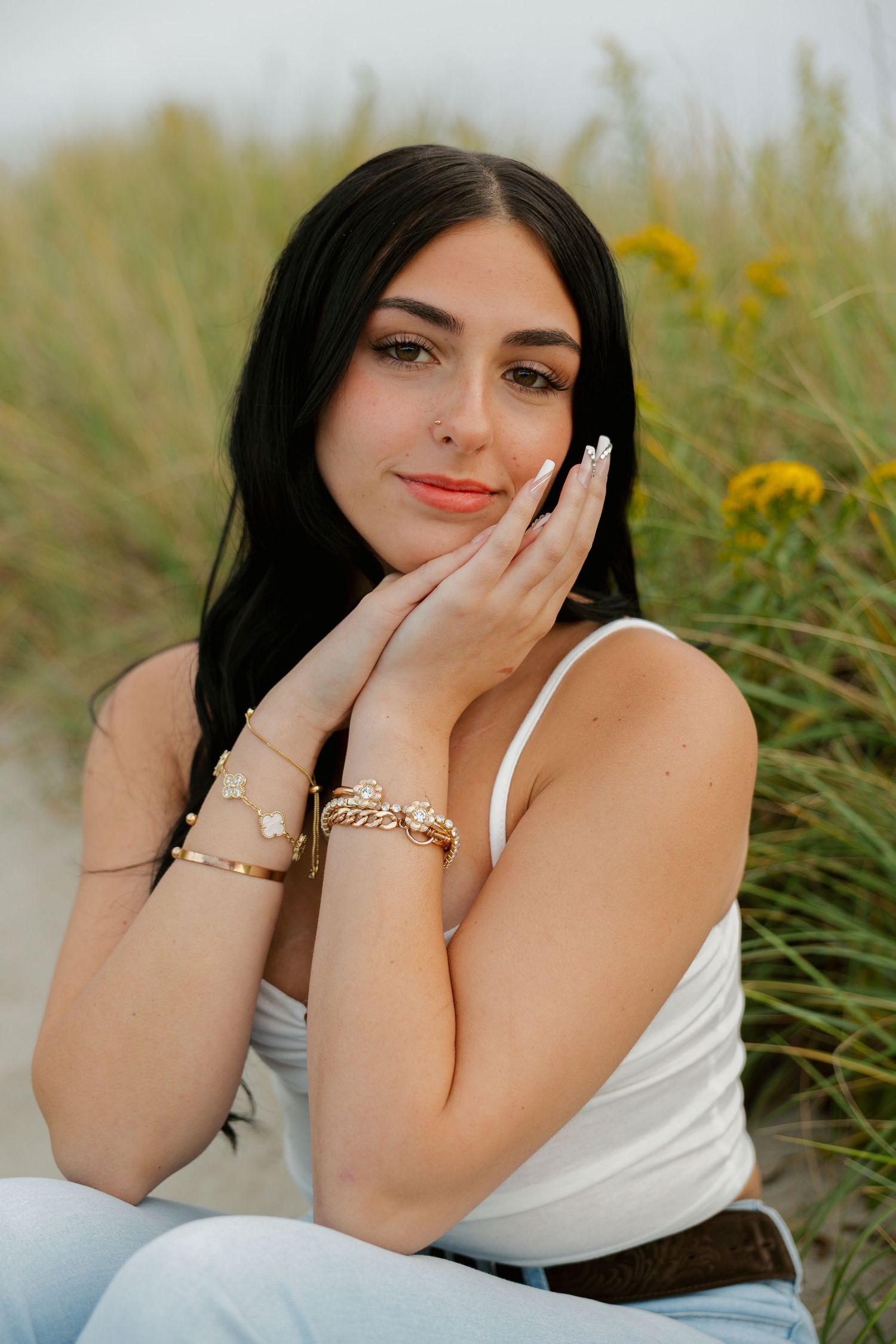 Young woman with dark hair wearing gold bracelets and white top poses gracefully outdoors near wildflowers.