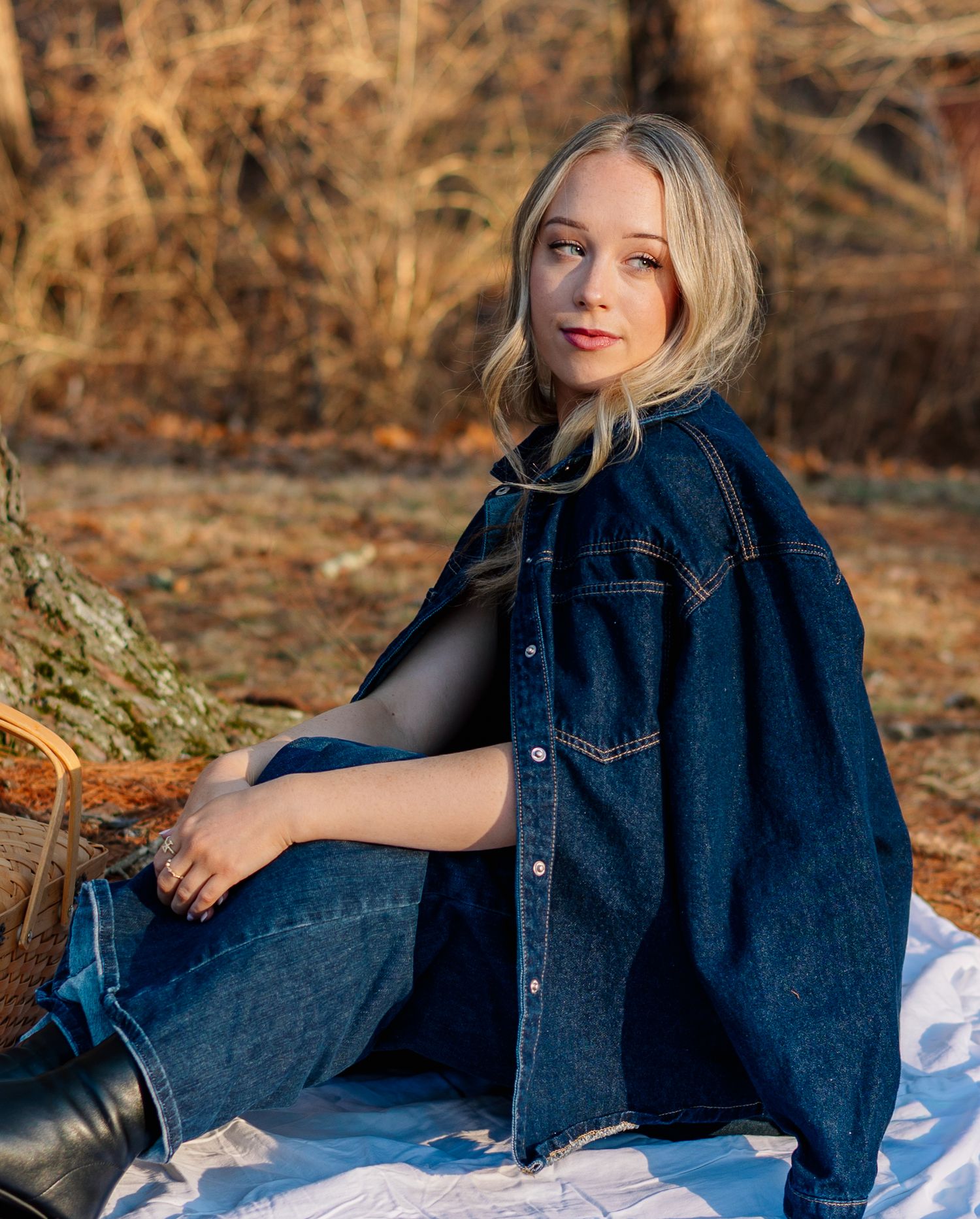 Blonde woman in dark denim jacket and jeans sitting outdoors on white blanket with picnic basket in autumn setting.