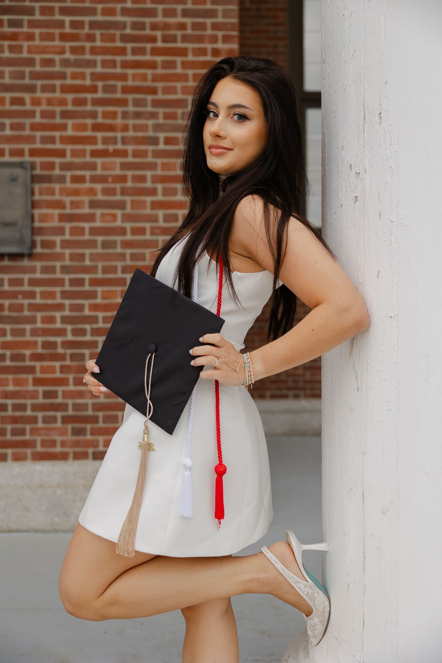 Young woman in white dress holding black graduation cap with red tassel, leaning against white column with brick wall background.