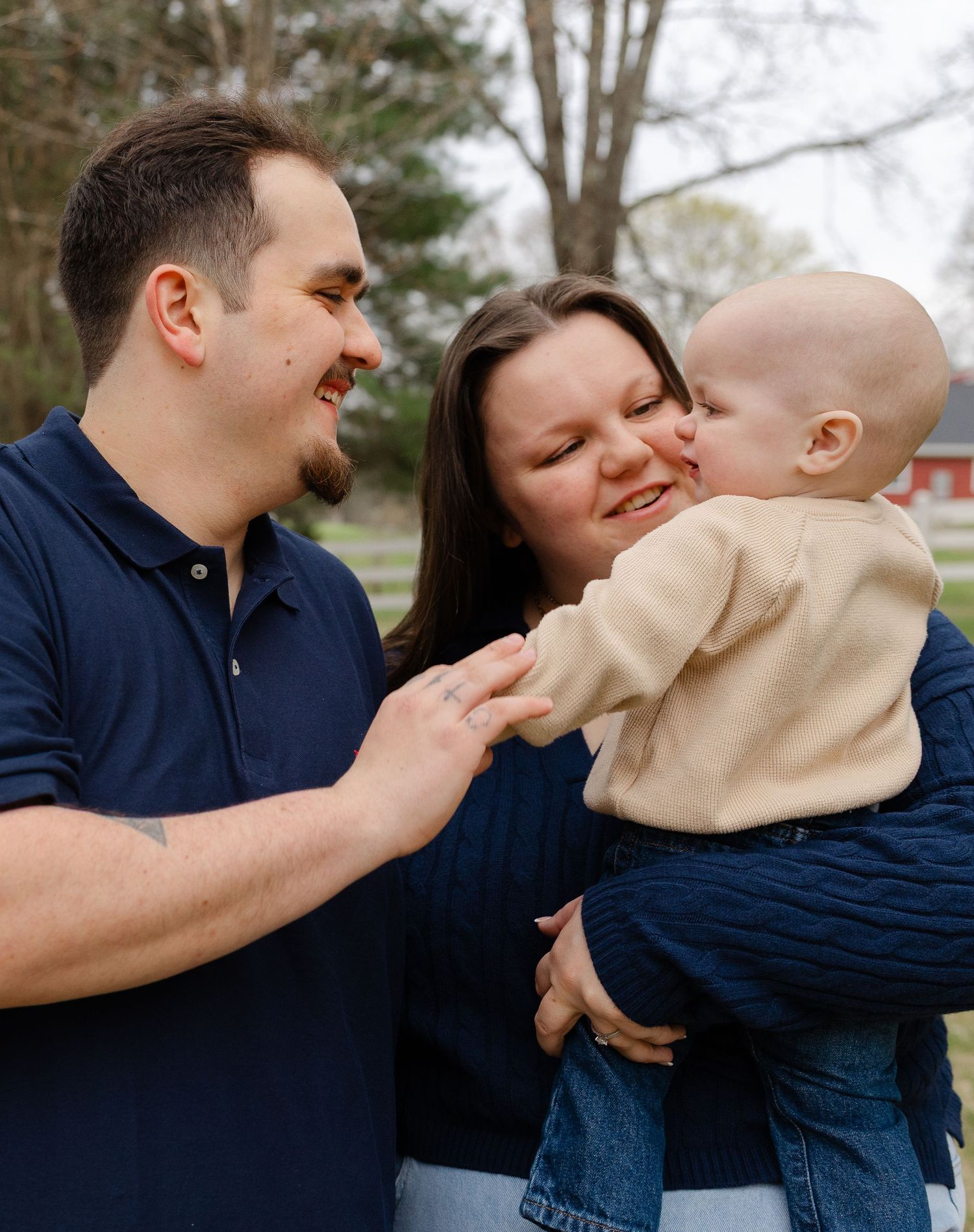 A young family smiles outdoors as a baby leans in to kiss the mother while the father looks on lovingly.