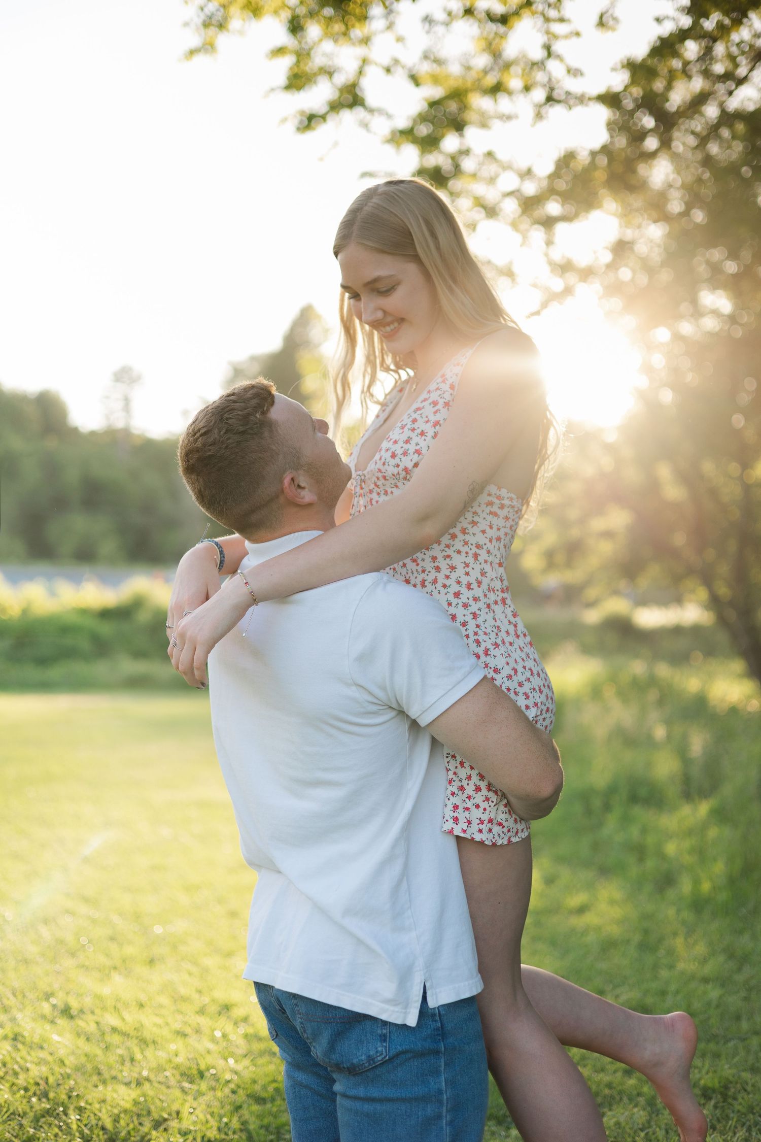 Man lifting smiling blonde woman in floral dress outdoors in glowing golden sunset park setting.