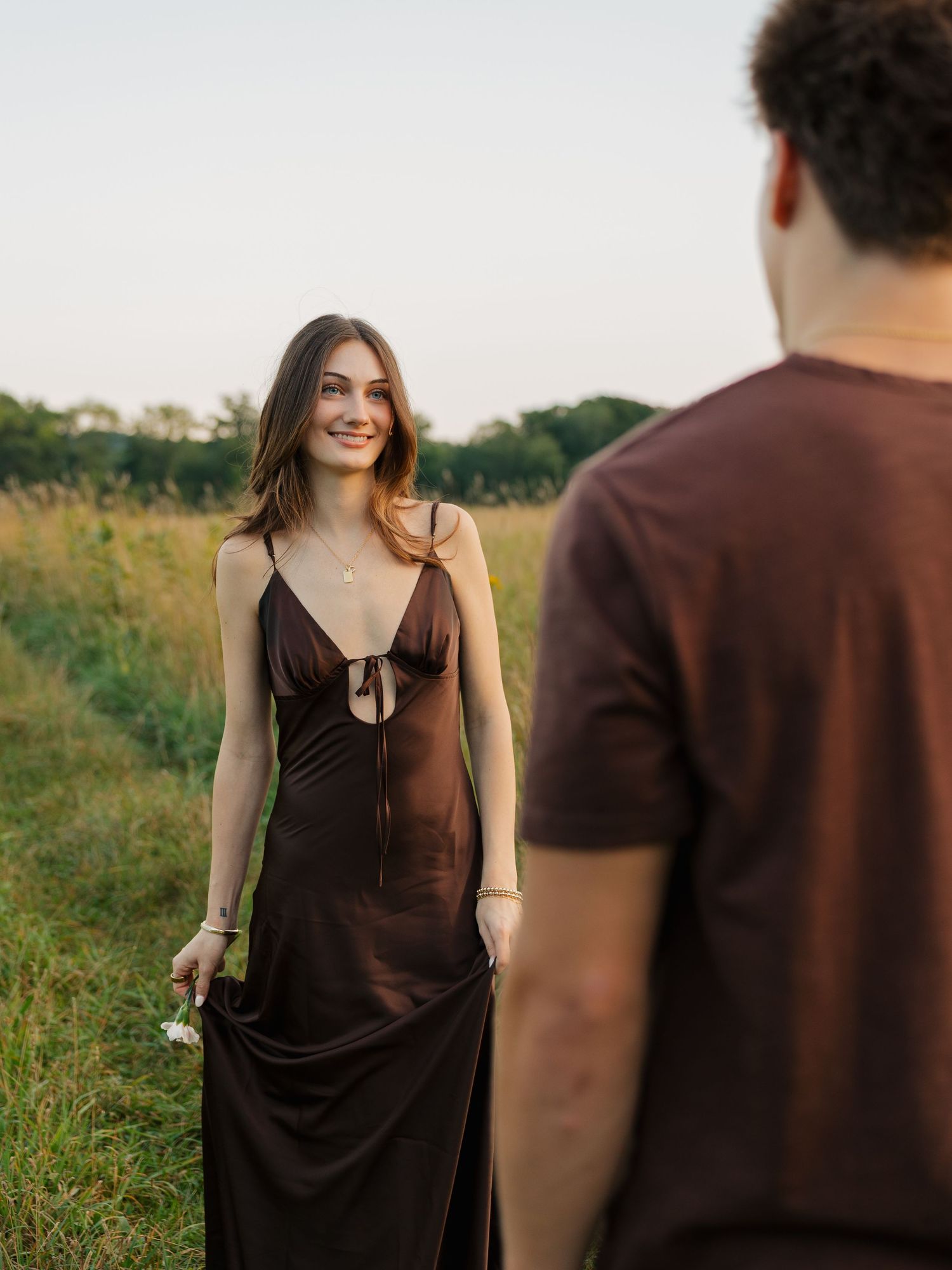 A woman in a brown satin slip dress smiles in a field, facing a man in a dark t-shirt.