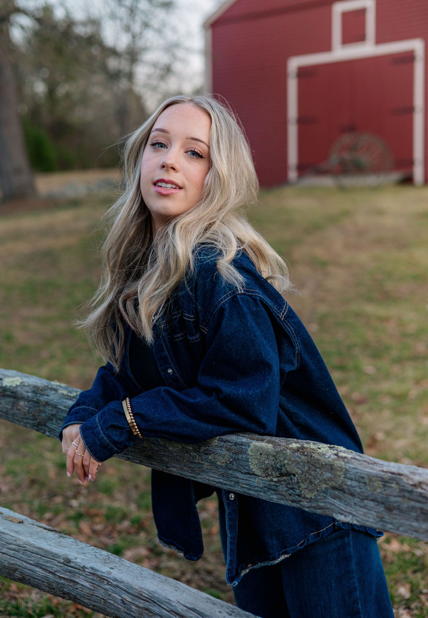 Blonde woman in dark denim jacket leaning on rustic wooden fence with red barn in background.