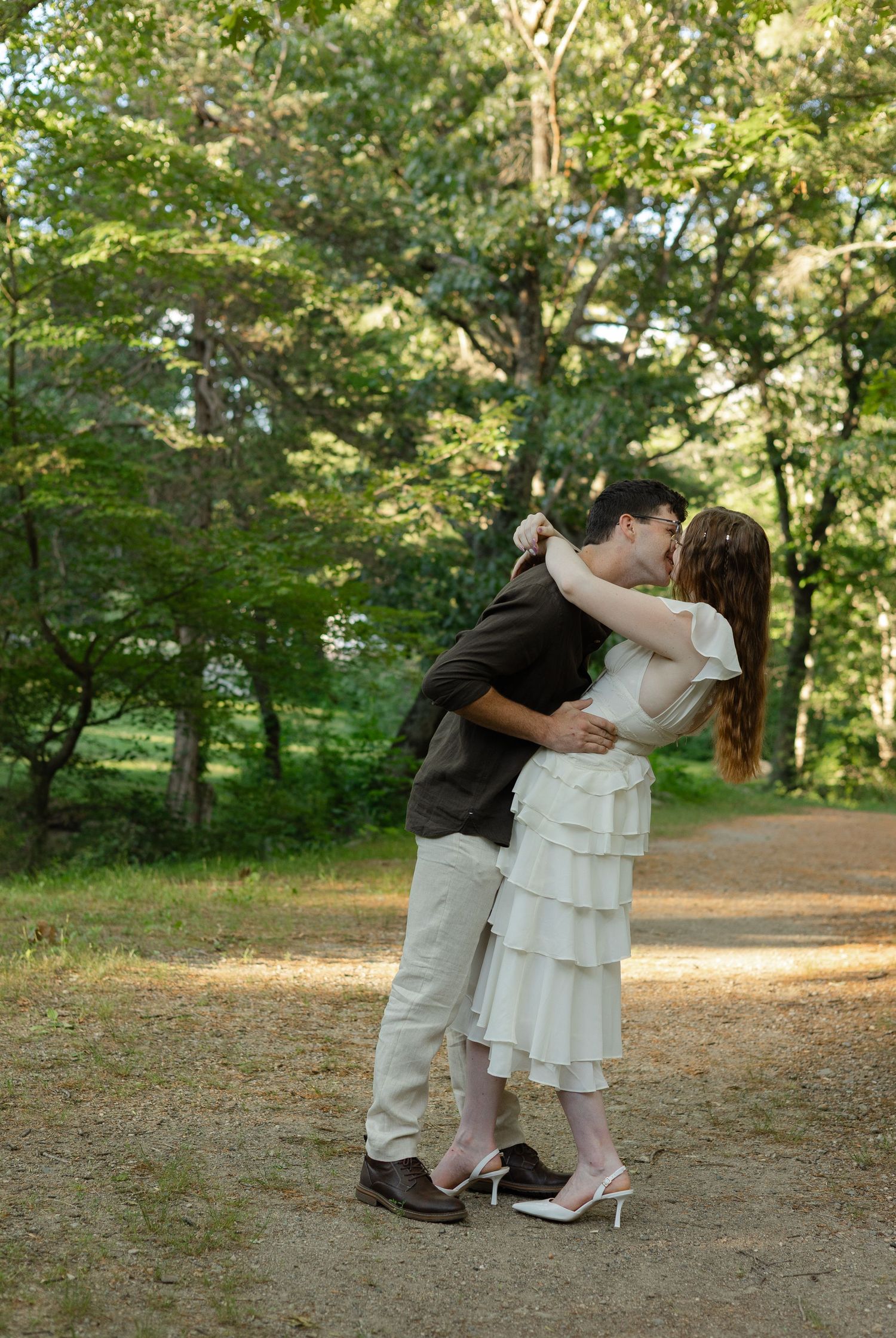 A couple shares a romantic kiss on a sunlit forest path, the woman wearing a white ruffled dress and heels.